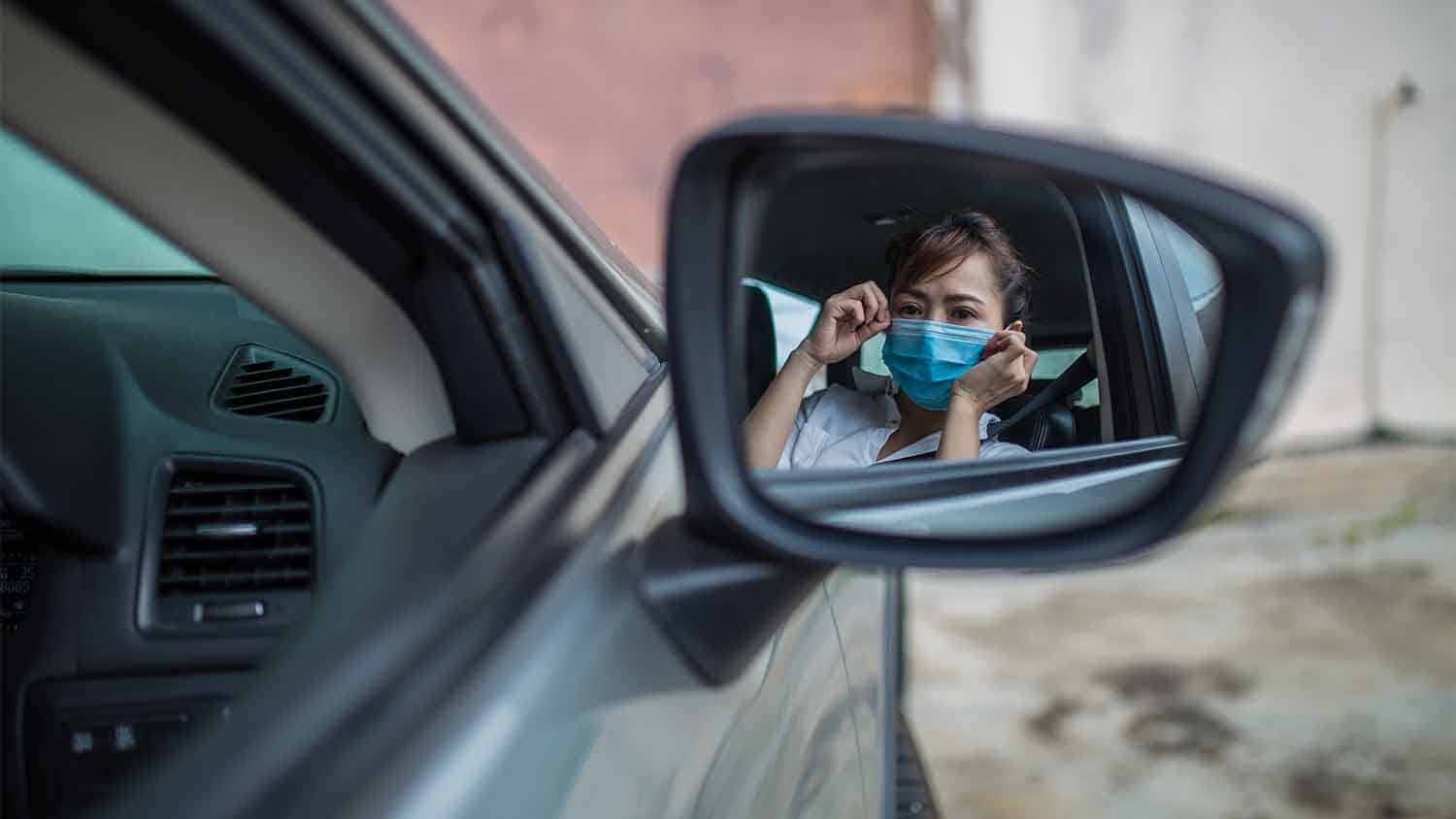 reflection of a woman putting on a face mask in a car wing mirror