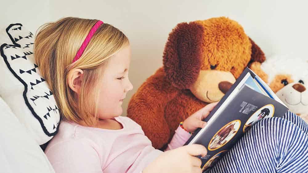 Little girl and a her teddy bear read a book.