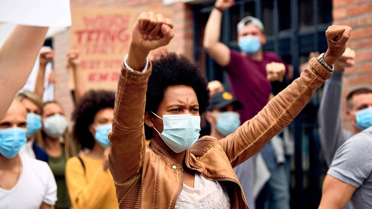 Black woman marching in protest arms aloft, ppe mask on