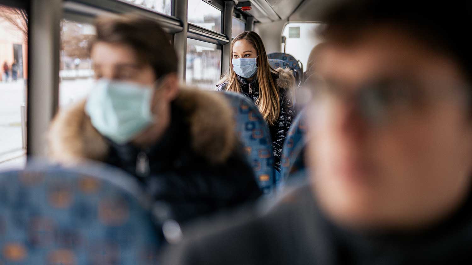 Passengers on bus, only the woman in the background is wearing a PPE mask