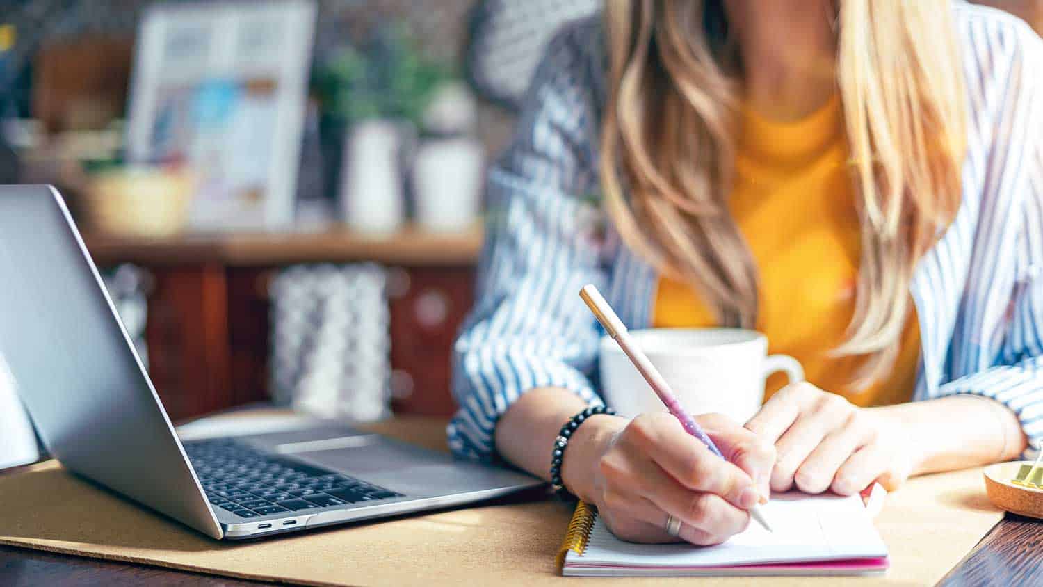 Woman working at a desk with a laptop and coffee. Pen is in hand writing on a pad