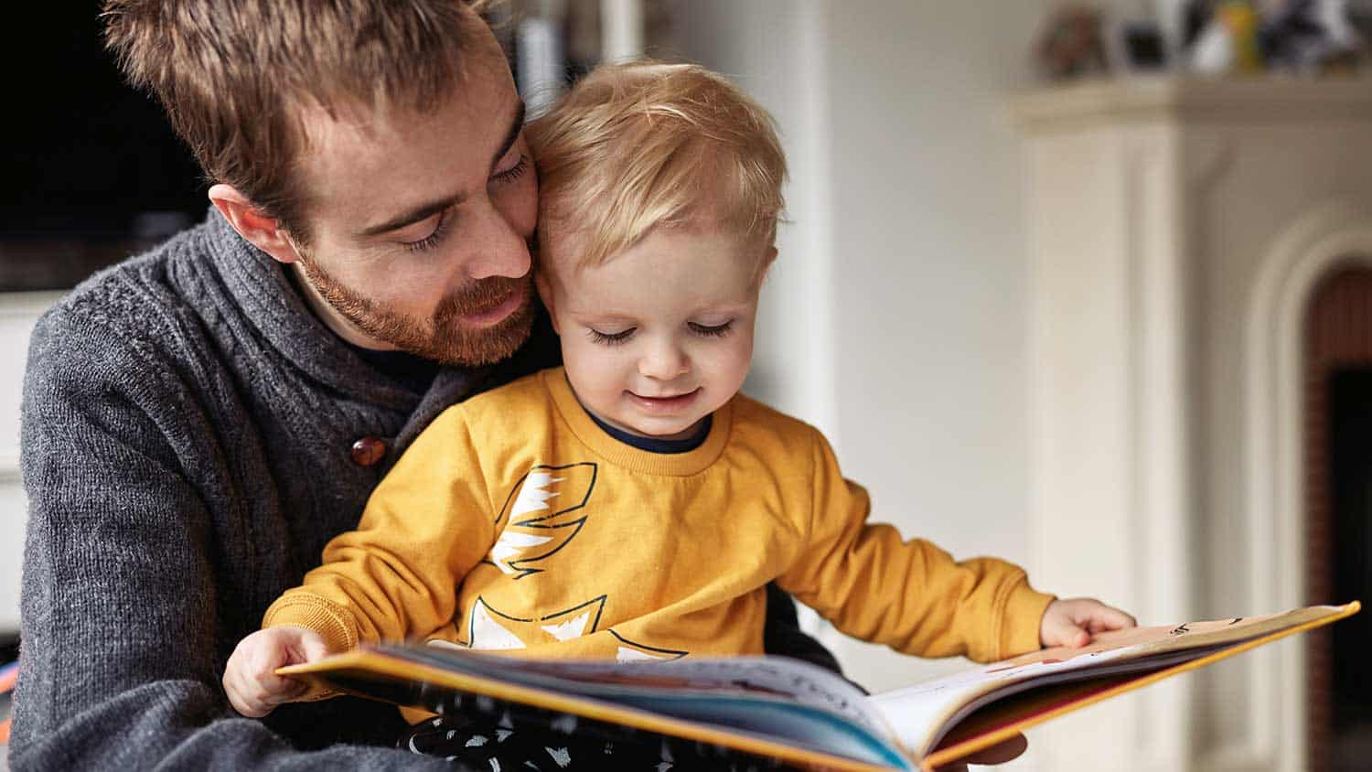 Child wearing a yellow jumper reading a story on his father's lap