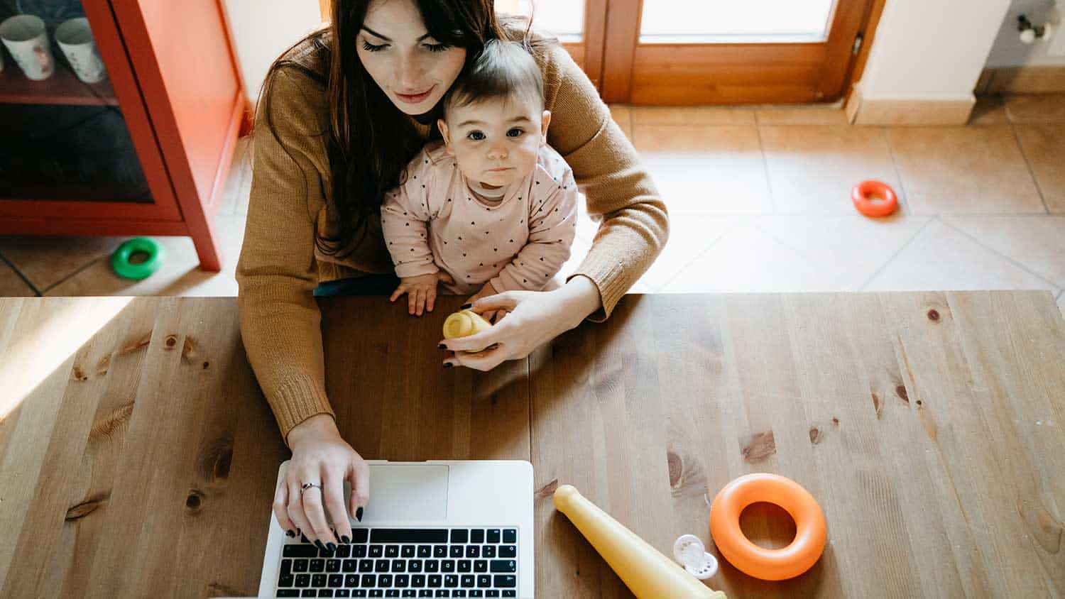 a mother working on a laptop holding a baby who is looking up to the camera