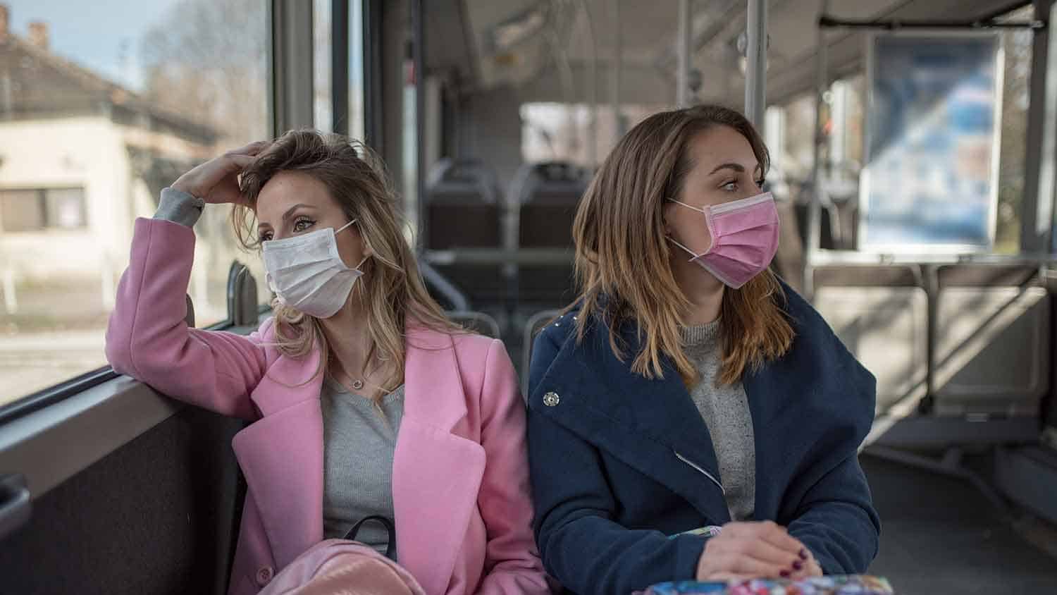Two women sat on a bus, both wearing face masks looking different directions