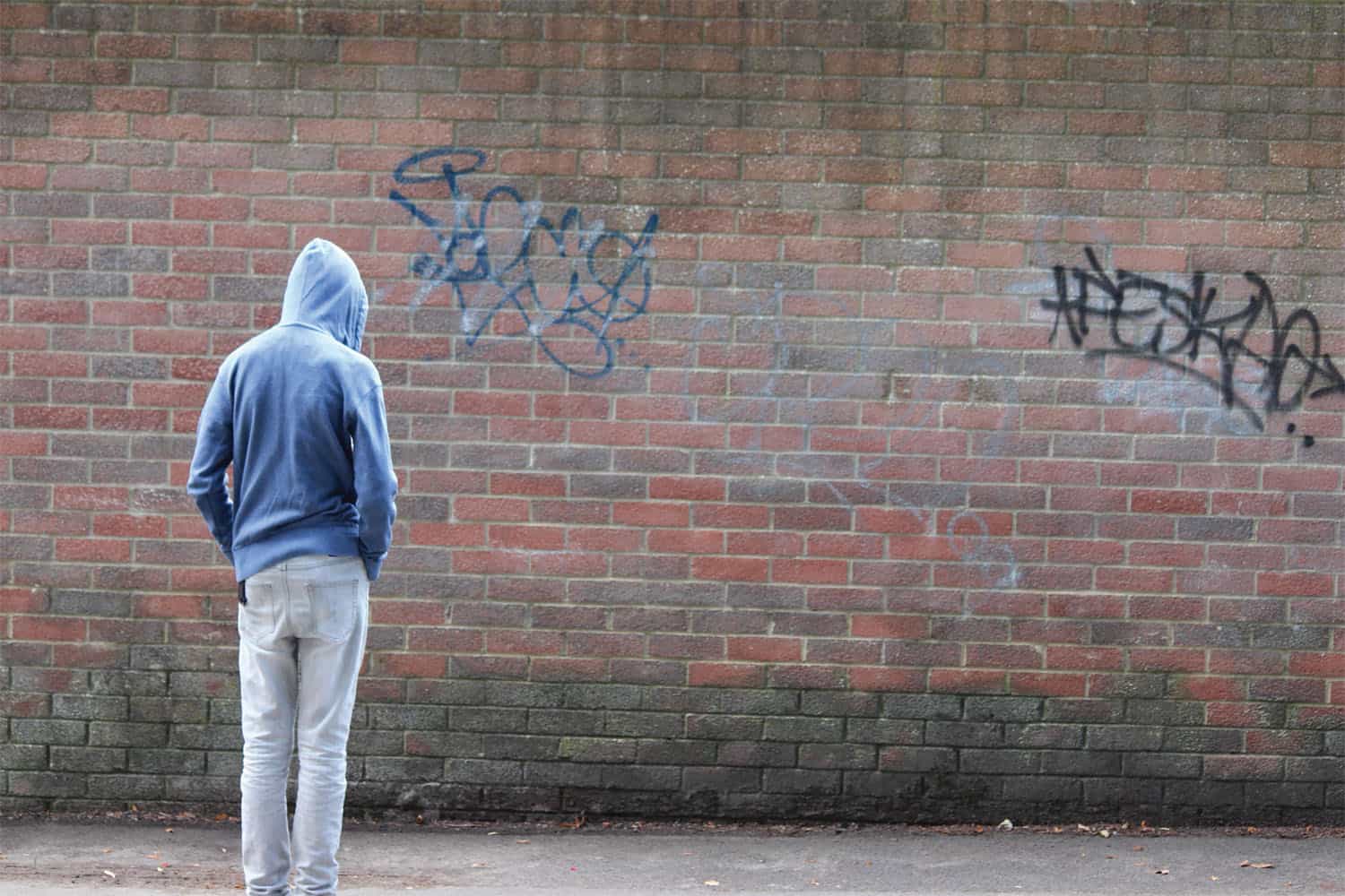Young person with their hood up facing a graffitied brick wall