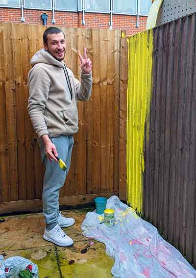 Man smiling at the camera doing a peace sign after painting a fence bright yellow