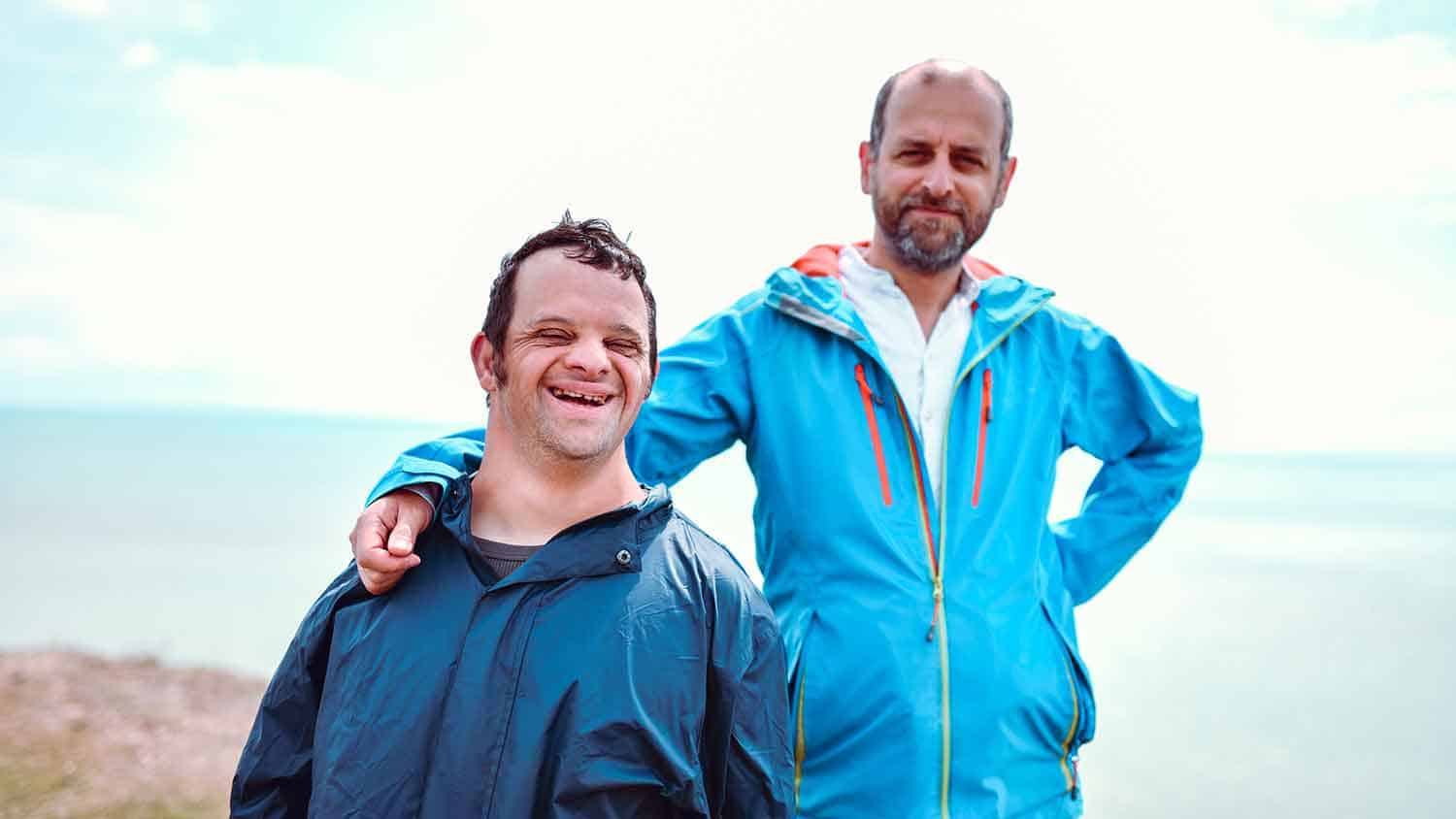 Two men looking at the camera in blue outdoor wear clothes. The man nearer the camera has down syndrome, the man behind has his hand on the other's shoulder