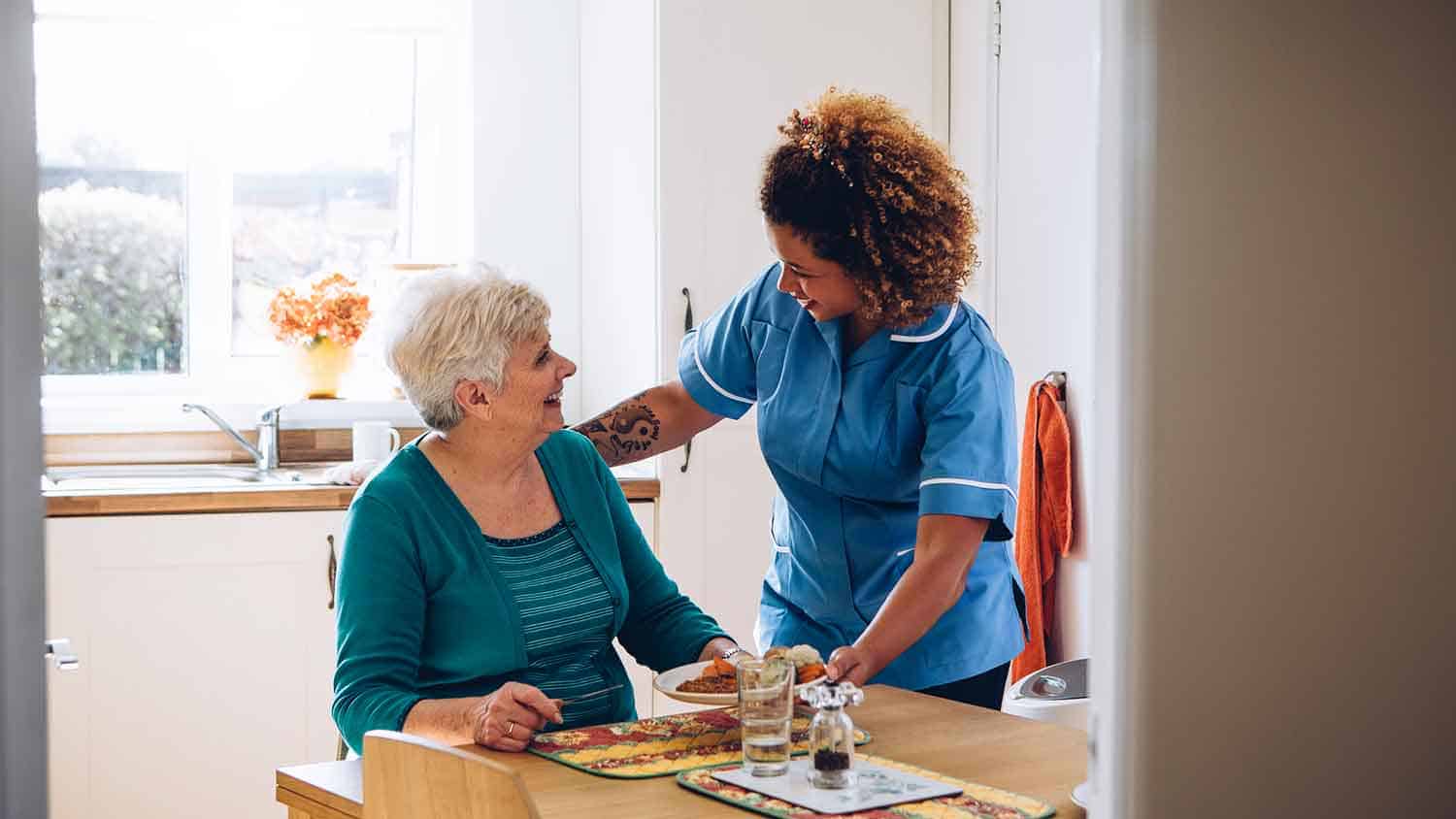 Nurse giving an older lady a meal. Both are smiling.