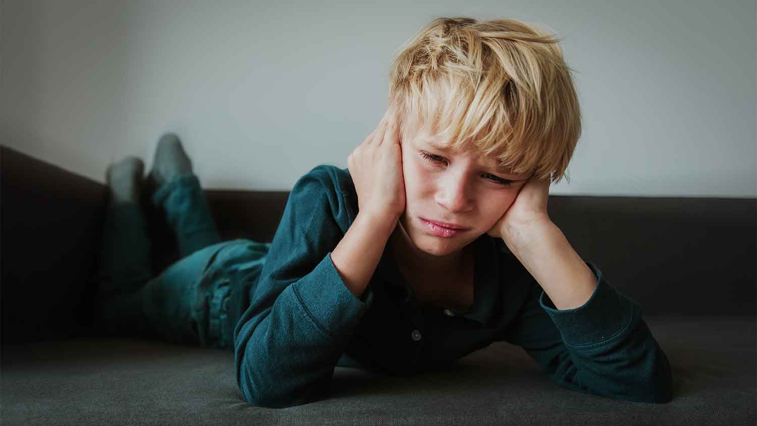 Young boy lying down with hands over his ears, looks sad.