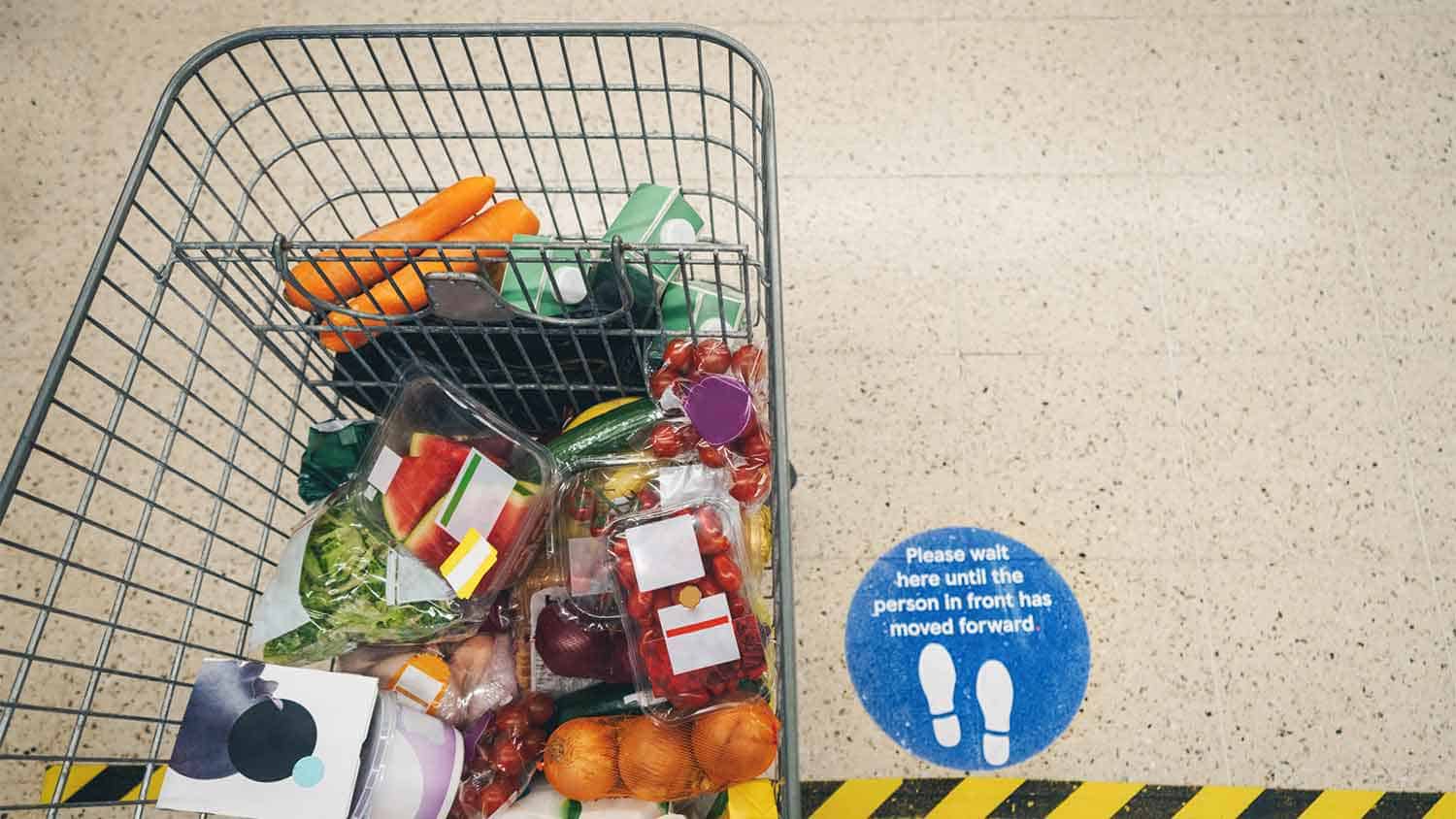 A supermarket trolley full of groceries. A covid 'keep your distance' sign is visible on the floor next to the trolley.