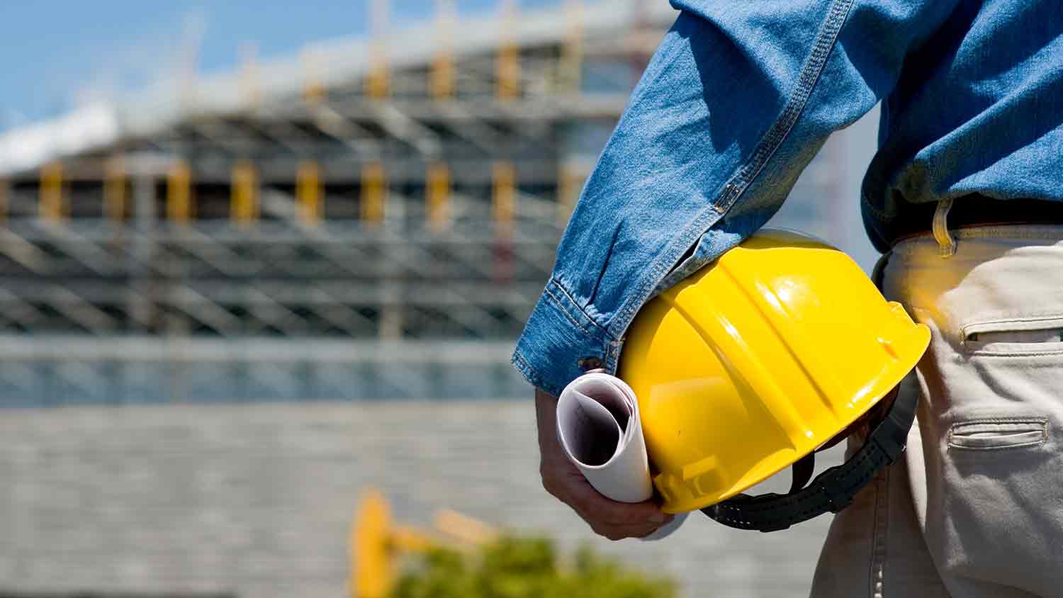 Person holdering a yellow hard hat and paper plans in front of a blurred construction site