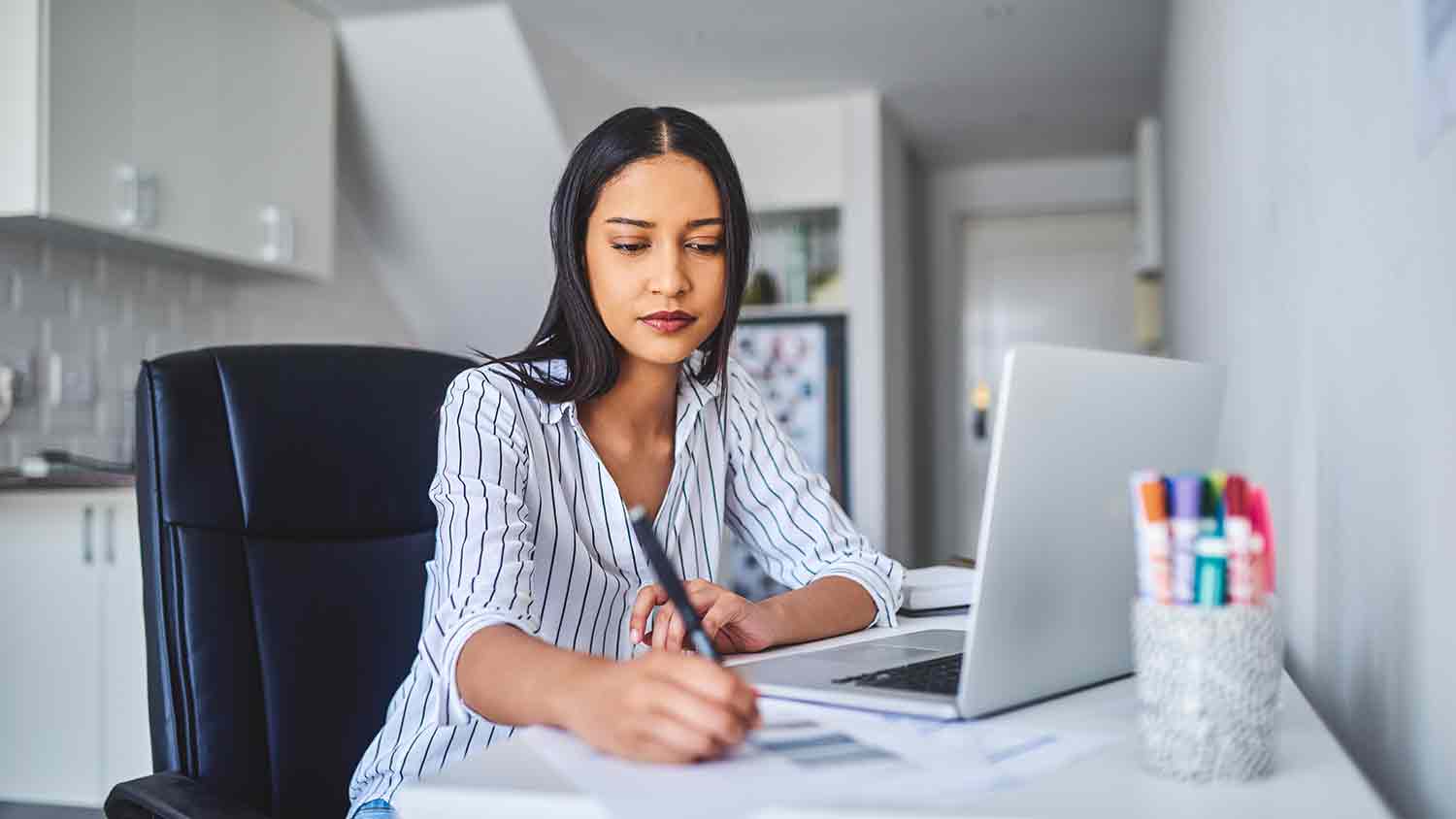 Working in a home office, based in a kitchen. Worker has a laptop and some coloured pens.