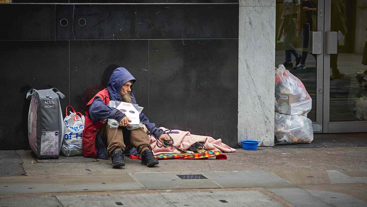 Homeless man with his belongings sat on a street. He is also holding onto a dog lead. His dog sits under a blanker next to him.