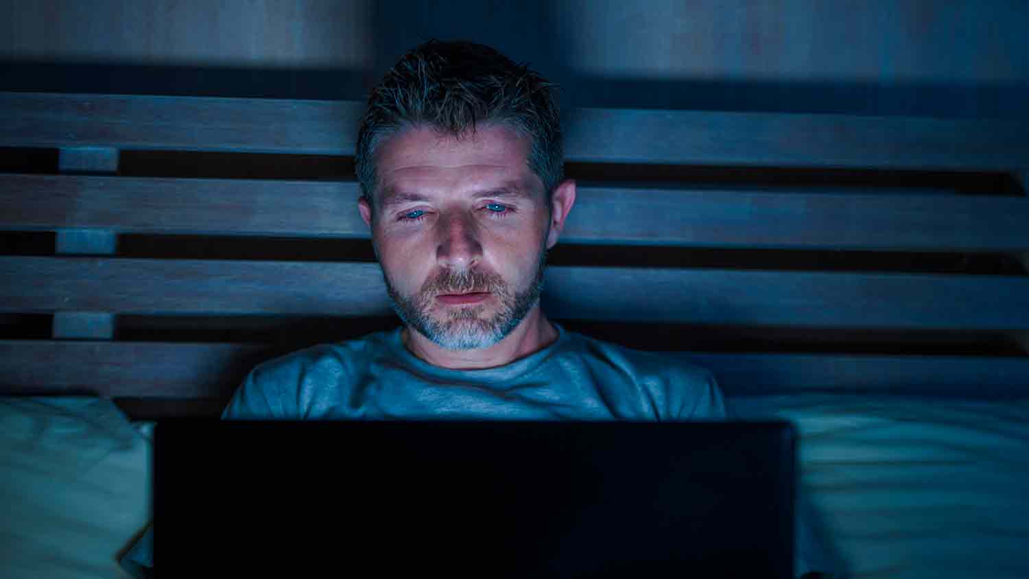 Man illuminated by a laptop in a dark bedroom. He appears upset.