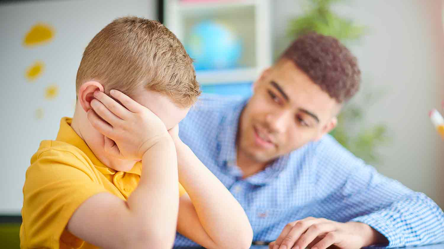 Young boy who is upset puts his hands over his eyes and ears not wanting to listen to a teacher who is leaning in