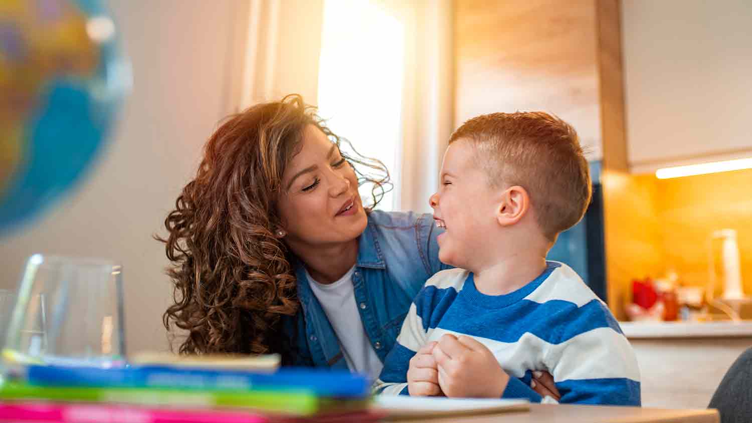 A young woman teaching likely her son at the kitchen table. Both are smiling and happy.