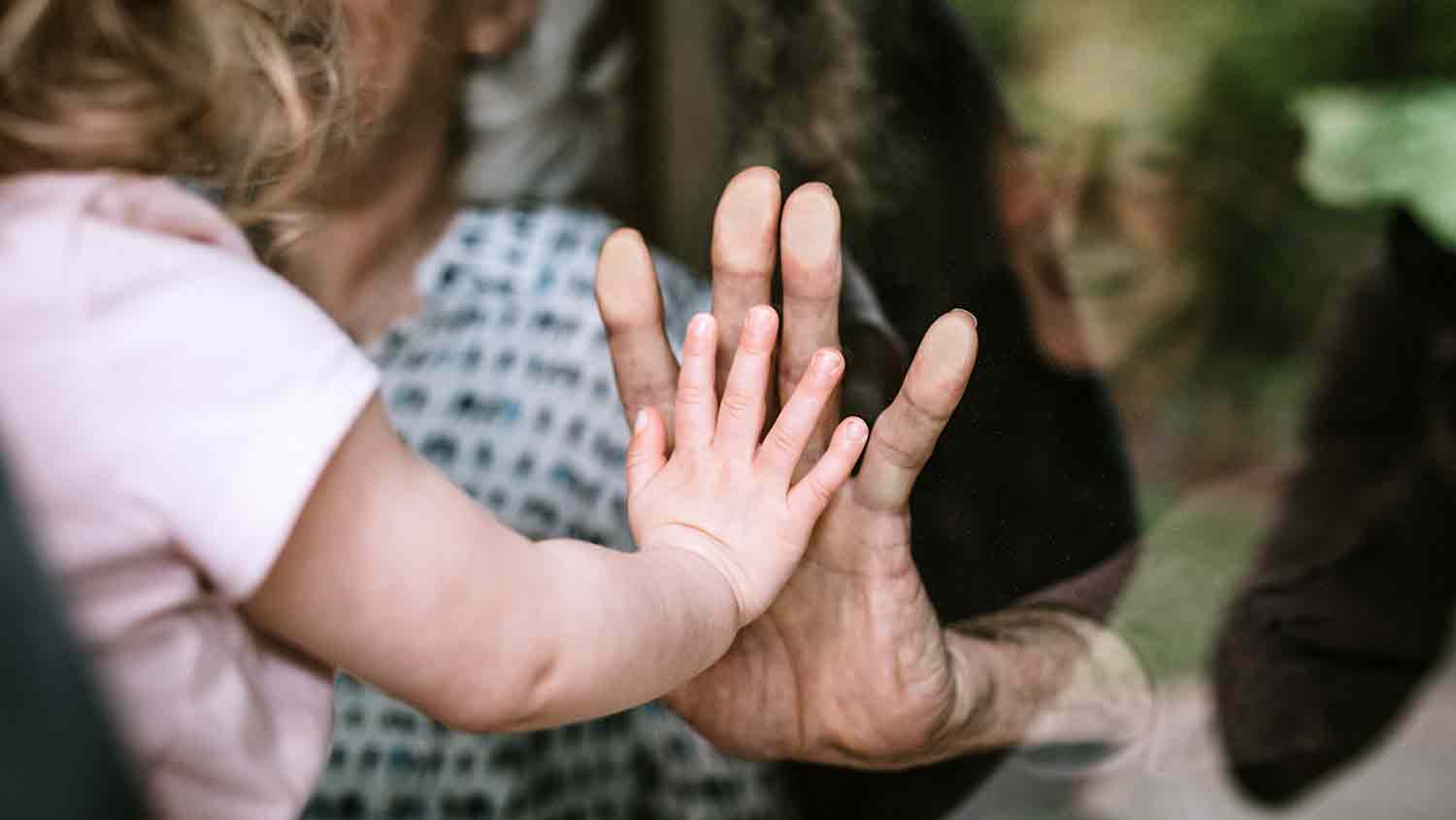 Toddler puts her hand to her granddad's. A pane of glass separates them.