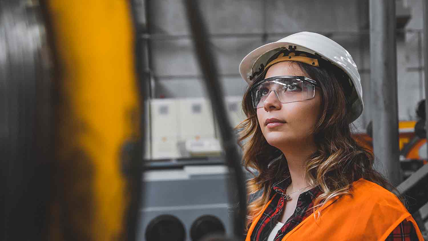Younug woman worker with high vis, safety glasses and white hard hat listens or observes