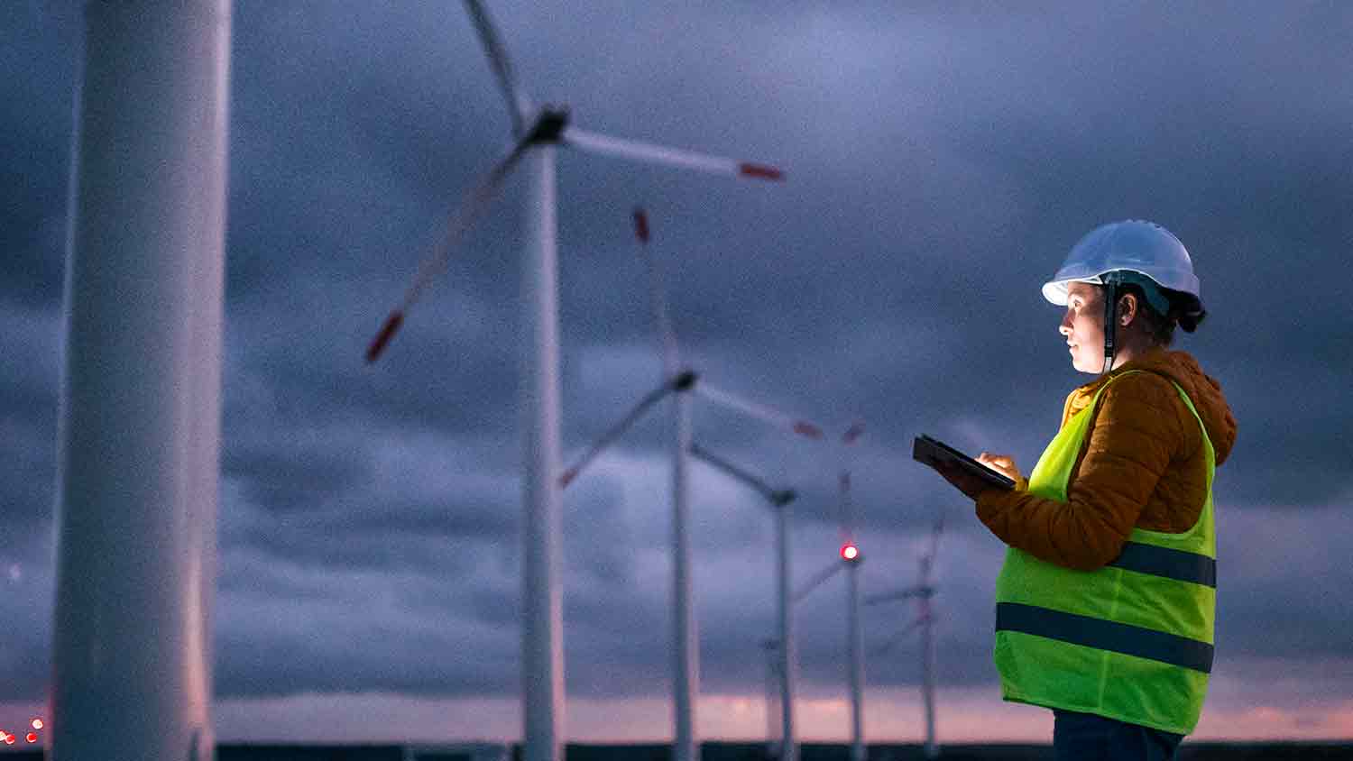 Engineer marking something on a tablet. The sky is dark and the tablet illuminates her face. The background is of wind turbines.