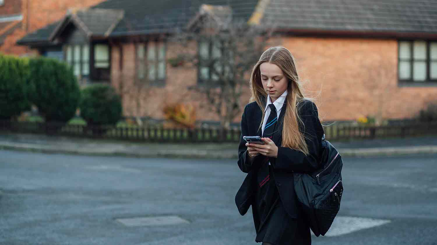 Teenage school girl walking in uniform across a road while scrolling on a mobile phone