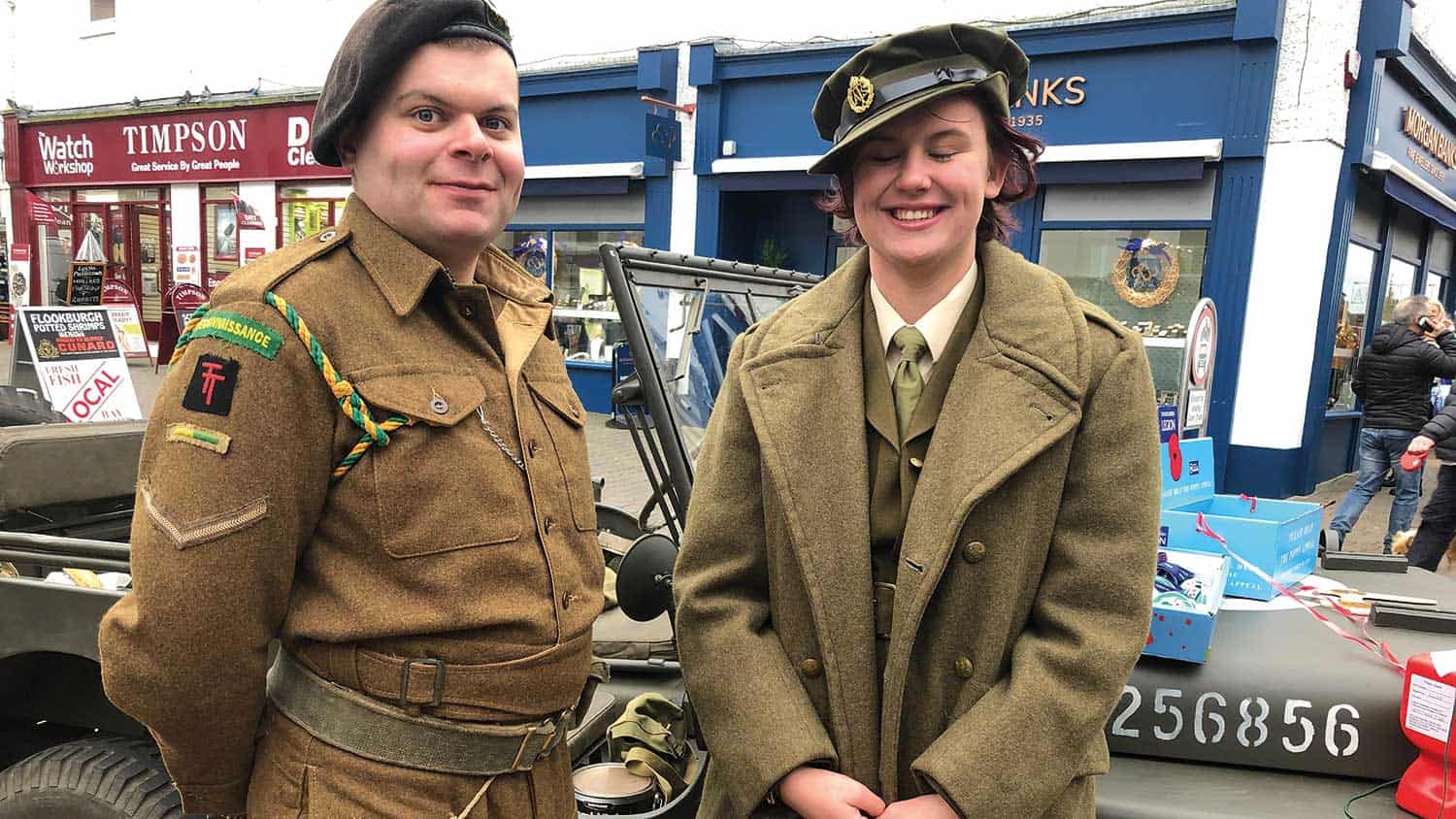 Two people stand in front of an old army vehicle, both wearing older style army uniforms