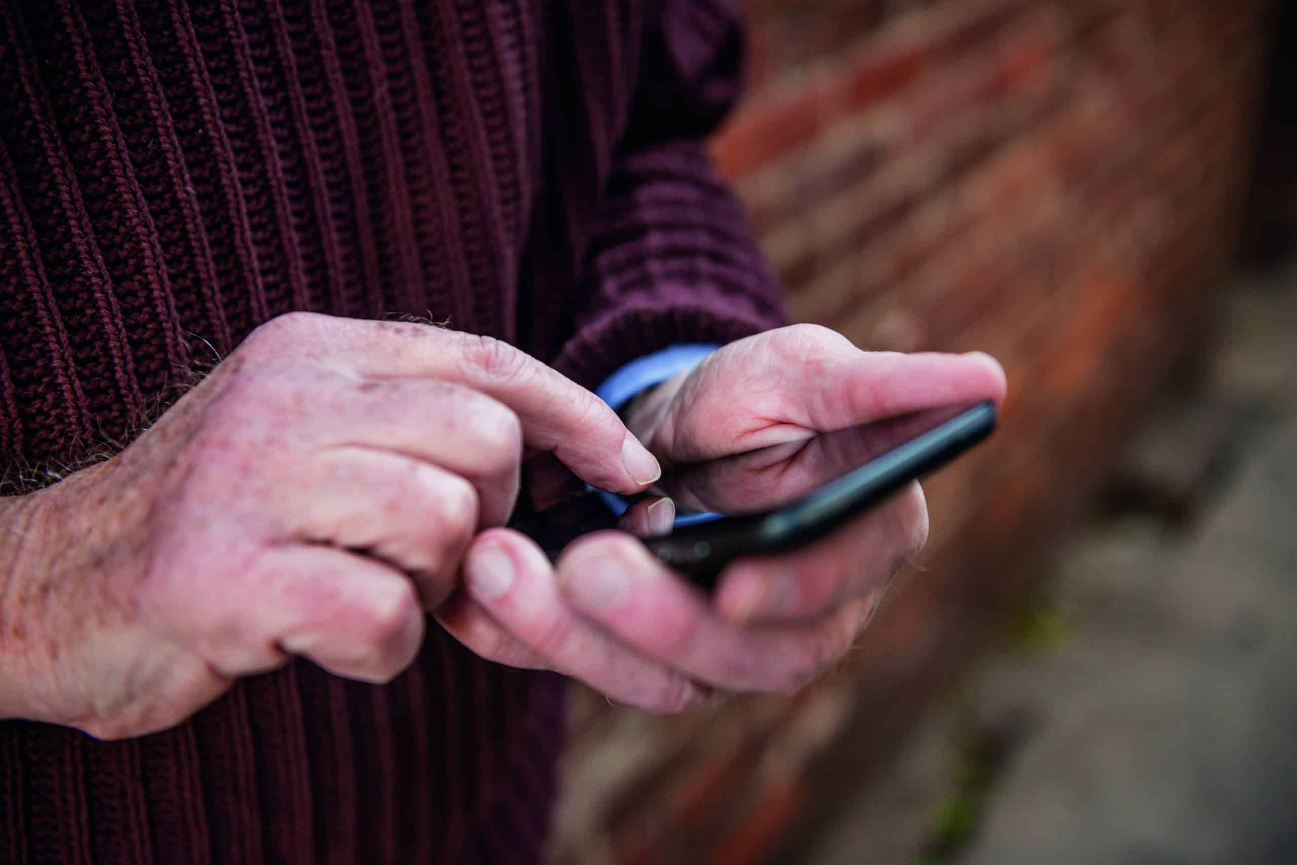 This is a close up color photograph of the hands of a senior Caucasian man in his 70s texting outdoors in New York City.