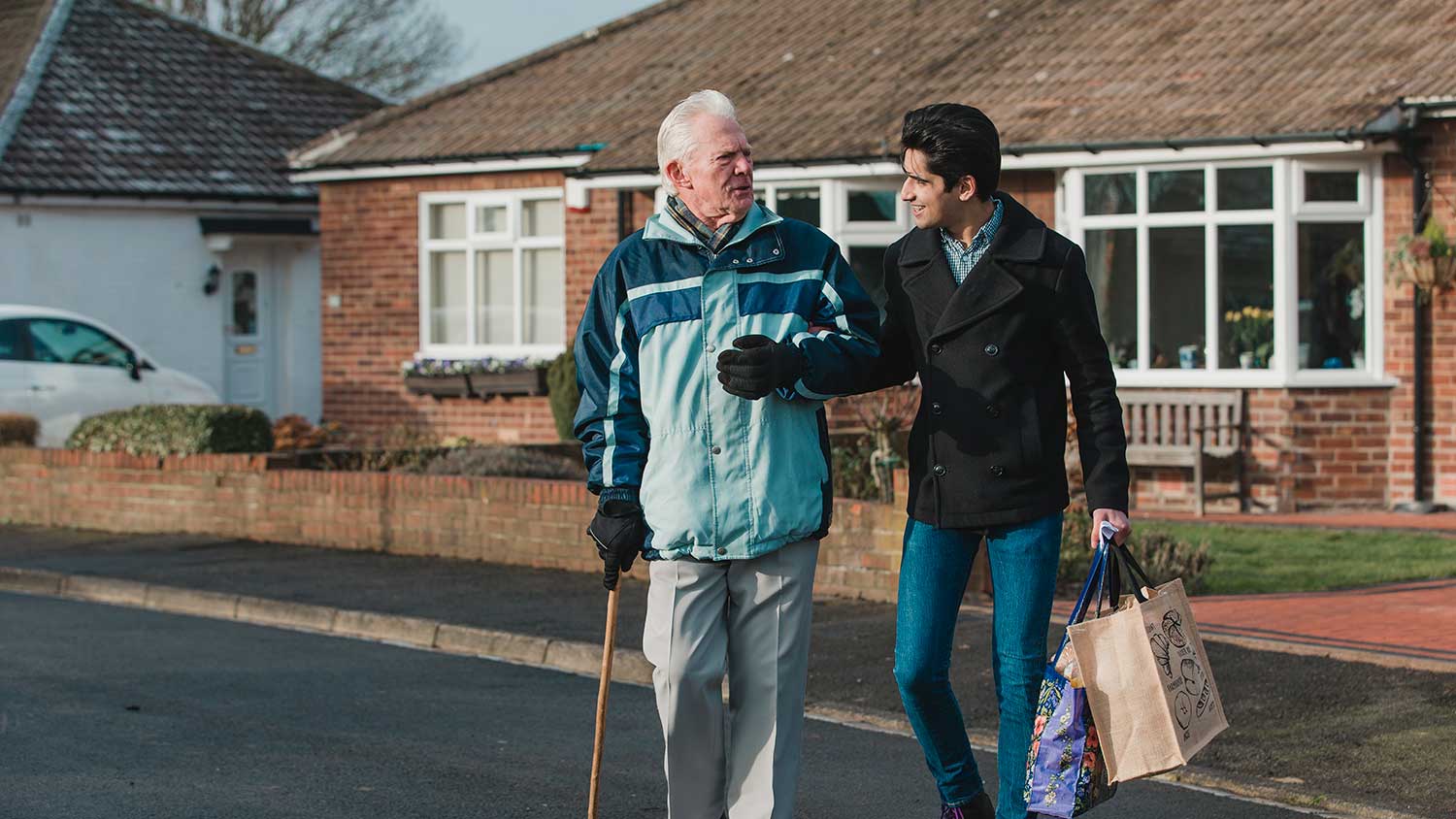 A younger man is helping an older man across the road. The younger man is carrying the shopping. The older is using a stick.