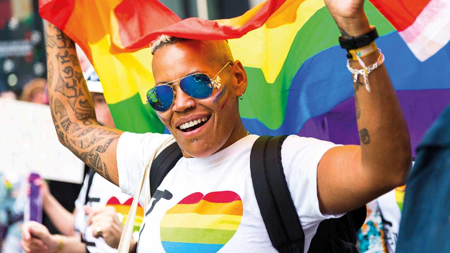 A person enjoying pride, holding a rainbow flag above their head and smiling to the camera