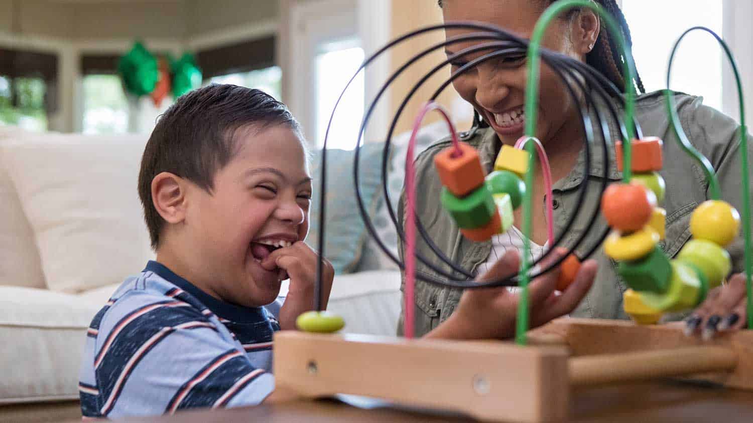 A child with down syndrome is playing with a toy, moving shapes across a wire. He and his teacher are laughing and happy.