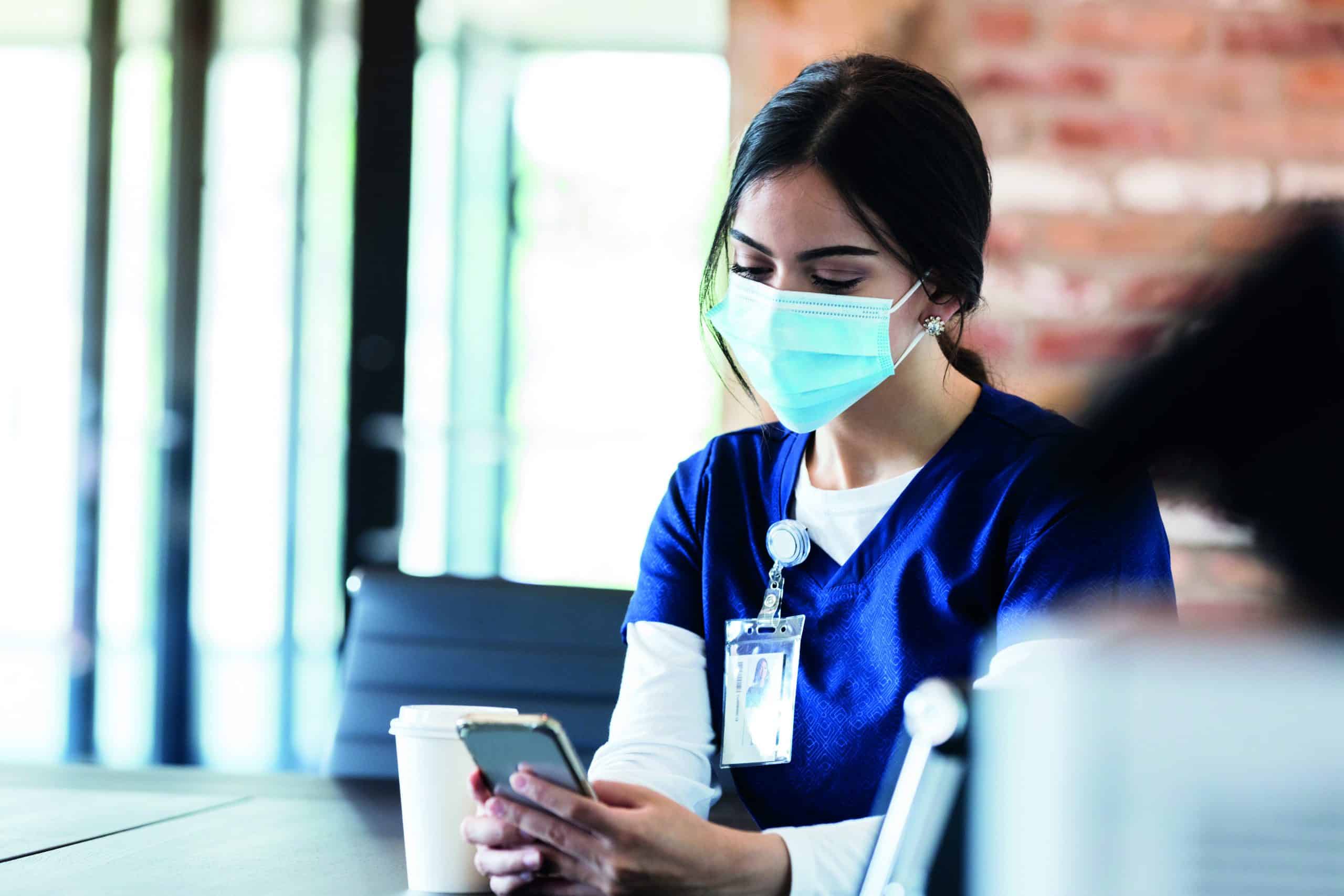 A female healthcare professional wears a mask while on break from her shift. She is checking for messages on her smartphone. She is wearing a protective face mask.