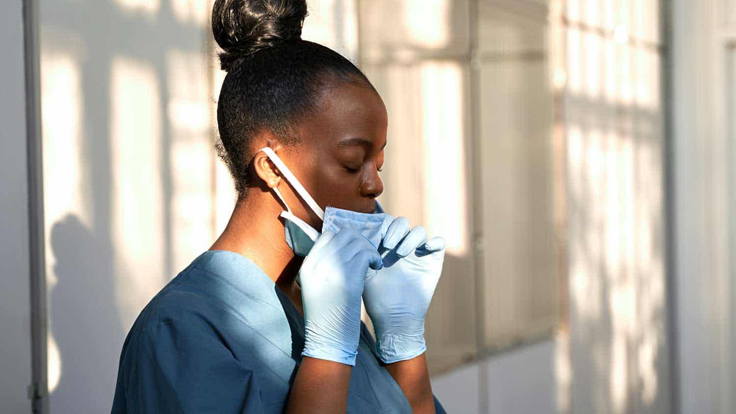 A nurse or doctor pulls down her PPE mask from her face. She's wearing all blue surgical gowns.