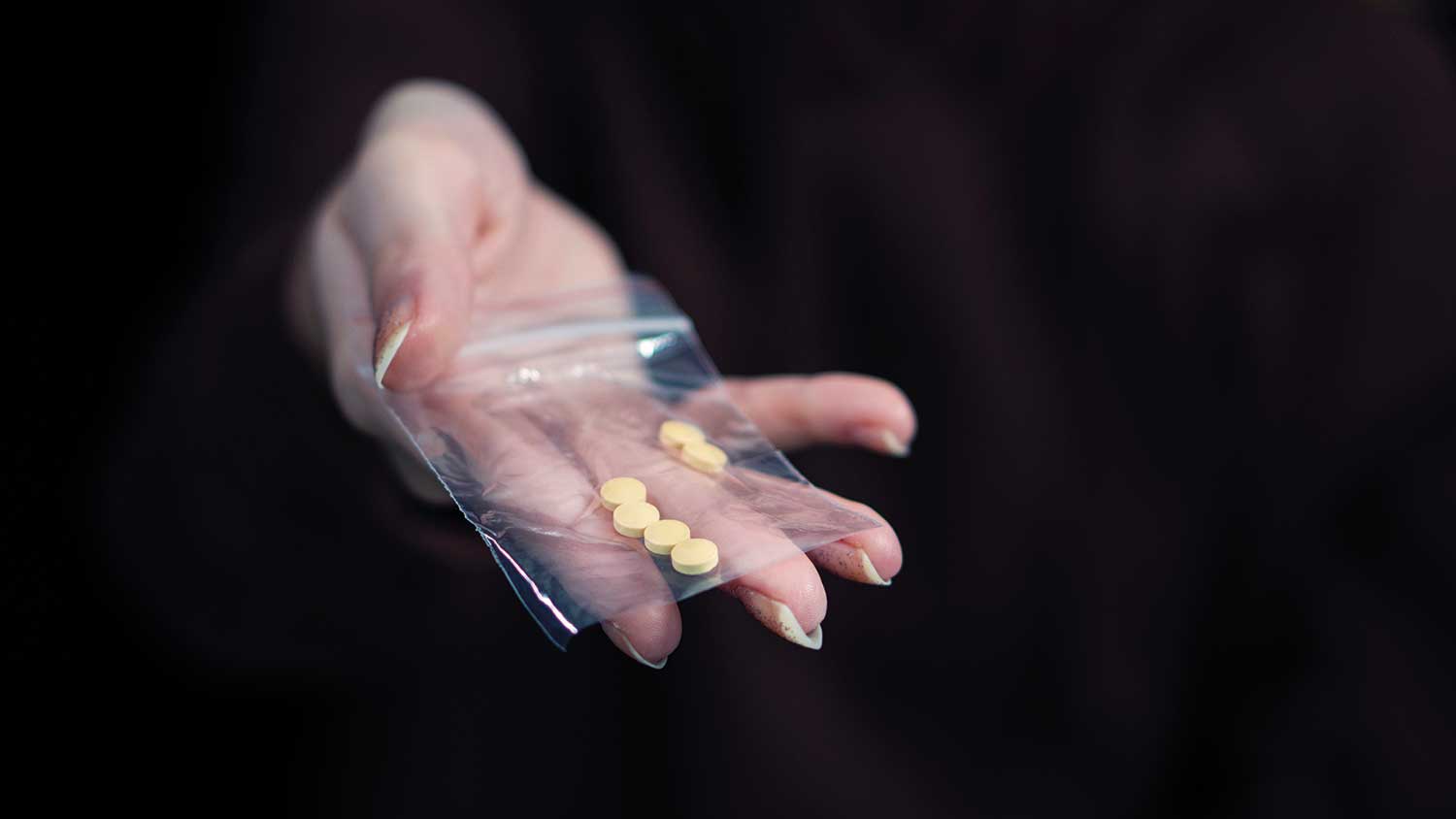 Six yellow pills in a clear plastic bag, held by a hand, the background is dark and all we see is the hand, bag and pills.