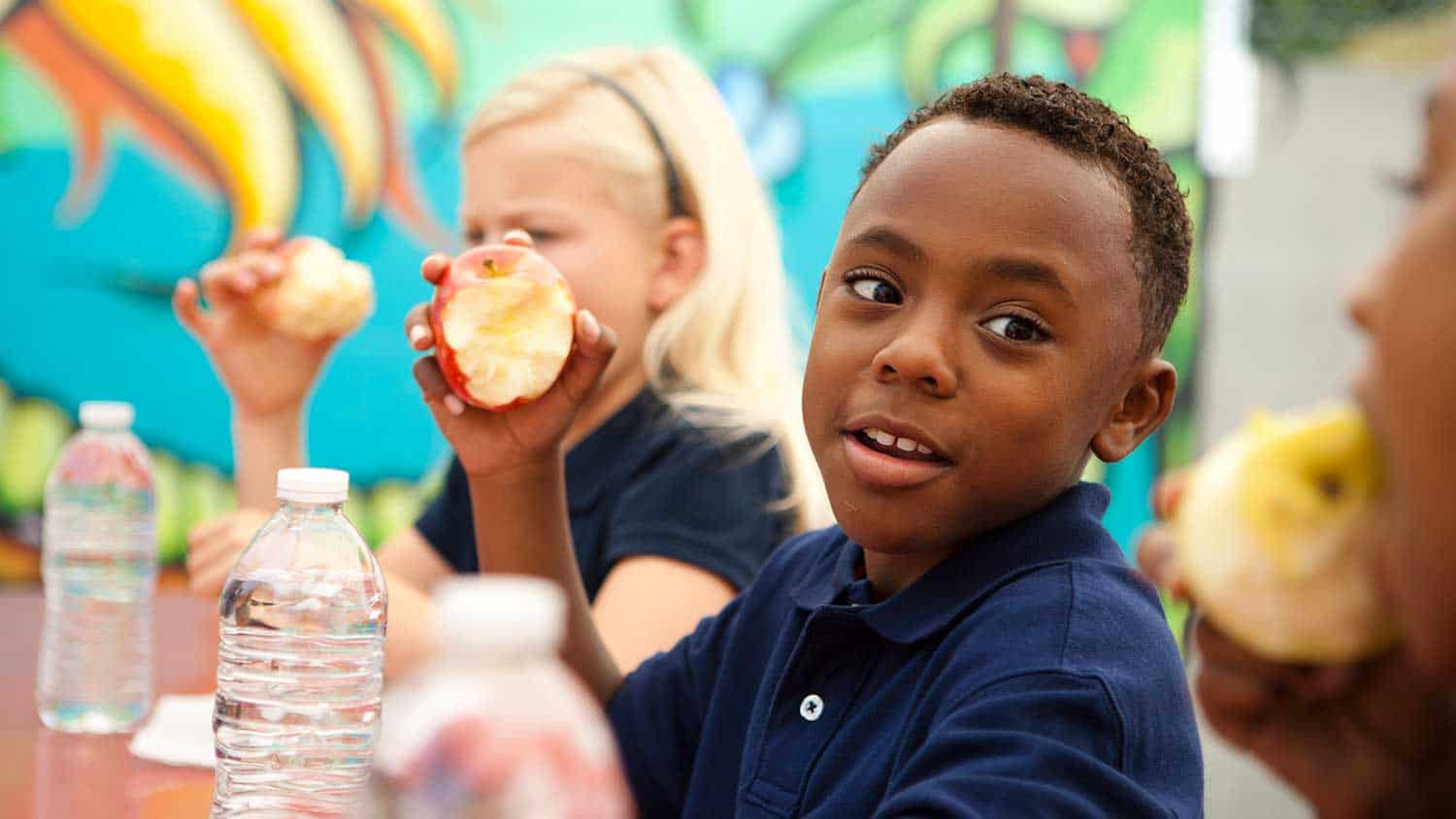 A young boy eating an apple as part of his packed lunch at school.