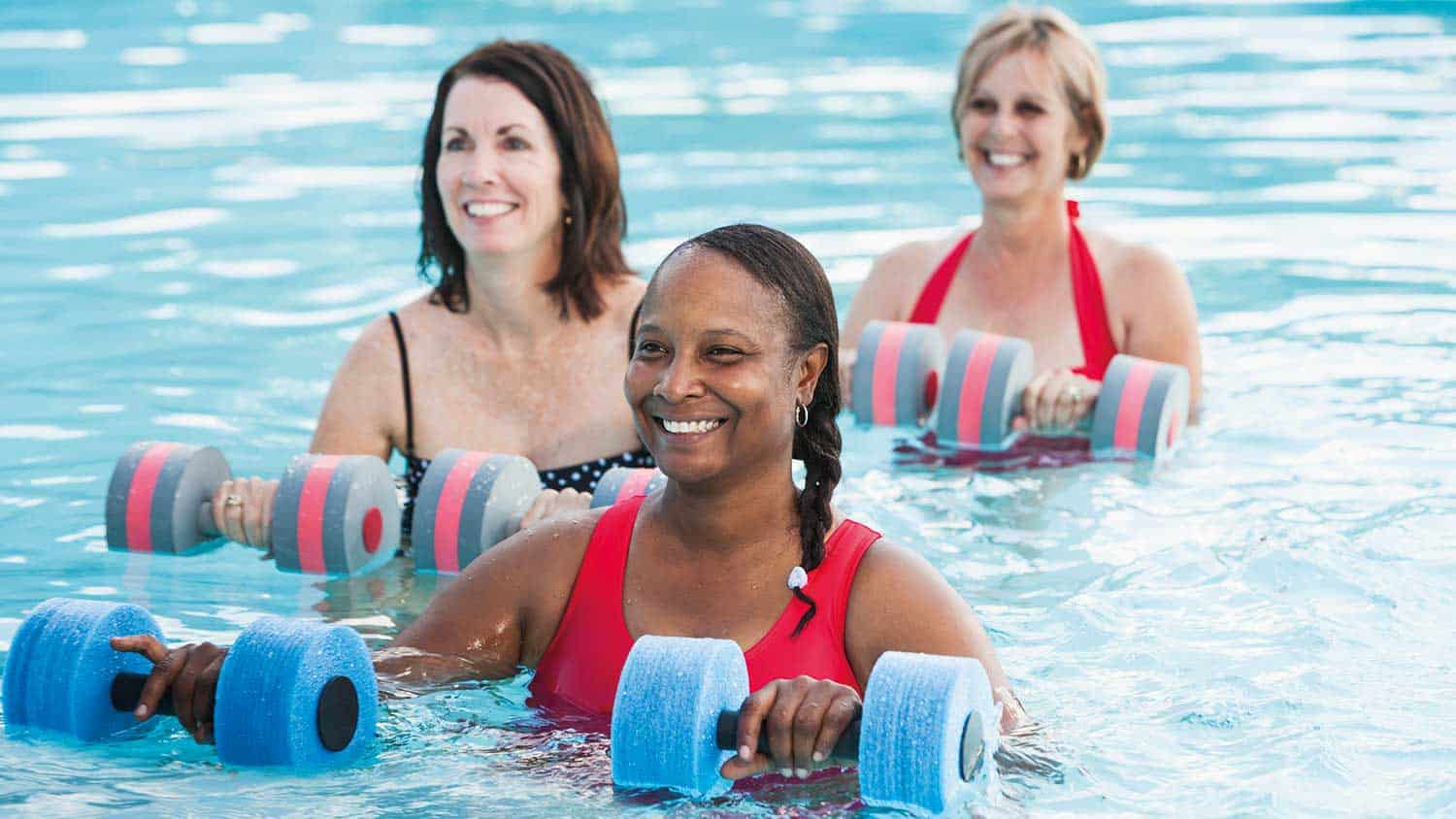 Three women looking at an instructor whilst standing in the water ready for an exercise class. They are holding water weights.