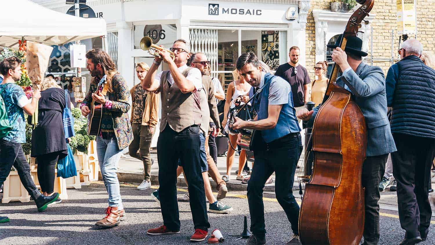 A jazz band plays in a street market. The trumpeter is blasting a note while the guitarist is concentrated.