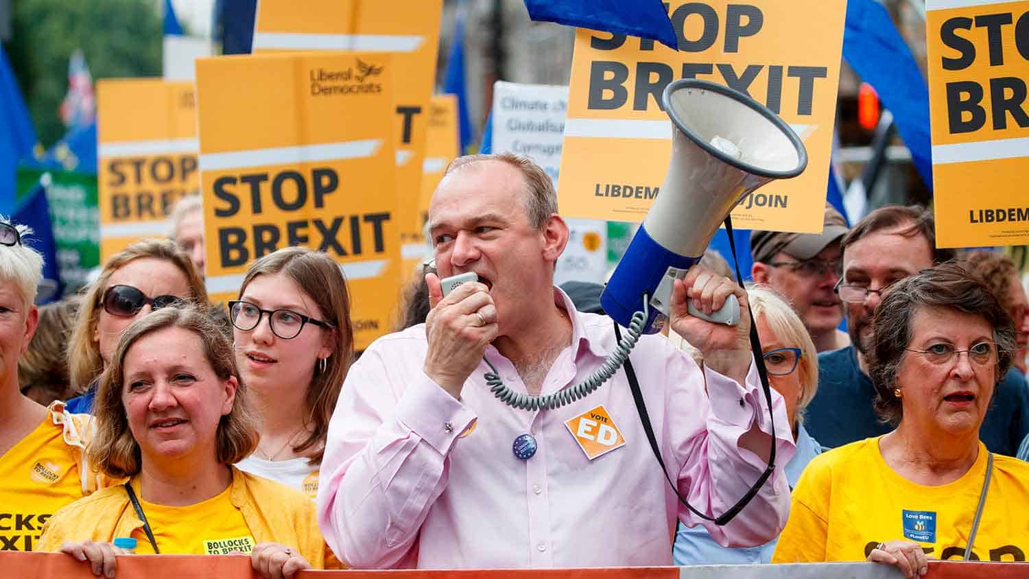 Sir Ed Davey MP addressing a crowd with a megaphone, in the background supporters are dressed in yellow