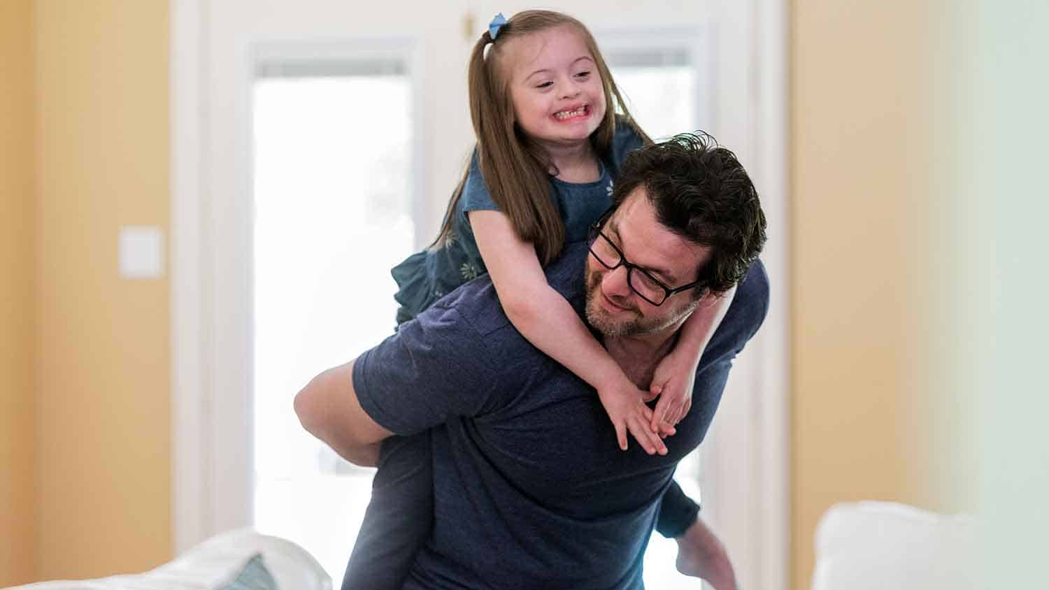 A young girl with Down Syndrome is given a piggy back by a man, both are laughing.