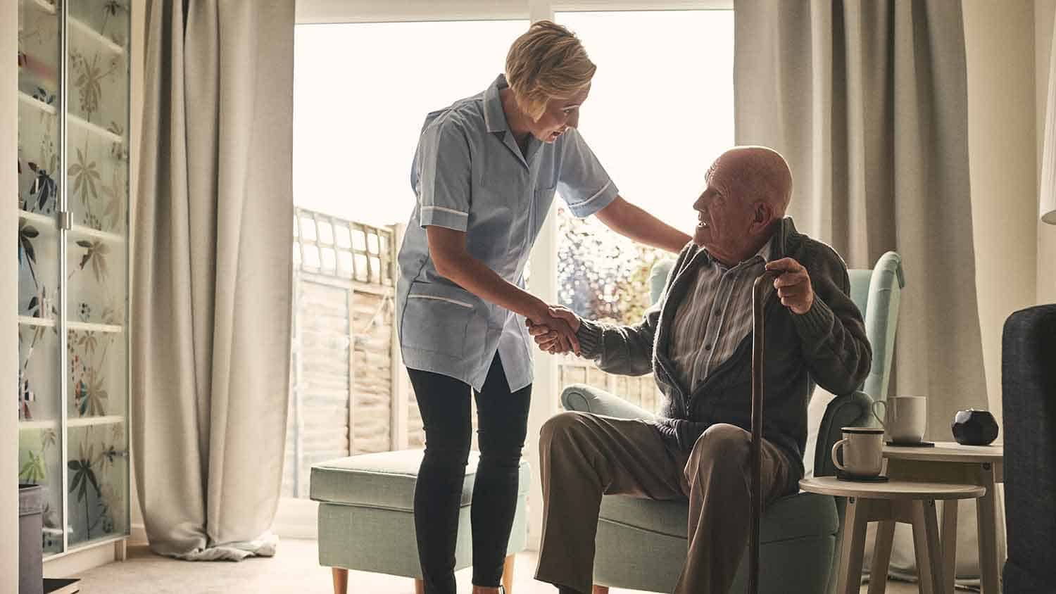 A carer and an older man holder hands. The older man in about to get up from a chair.