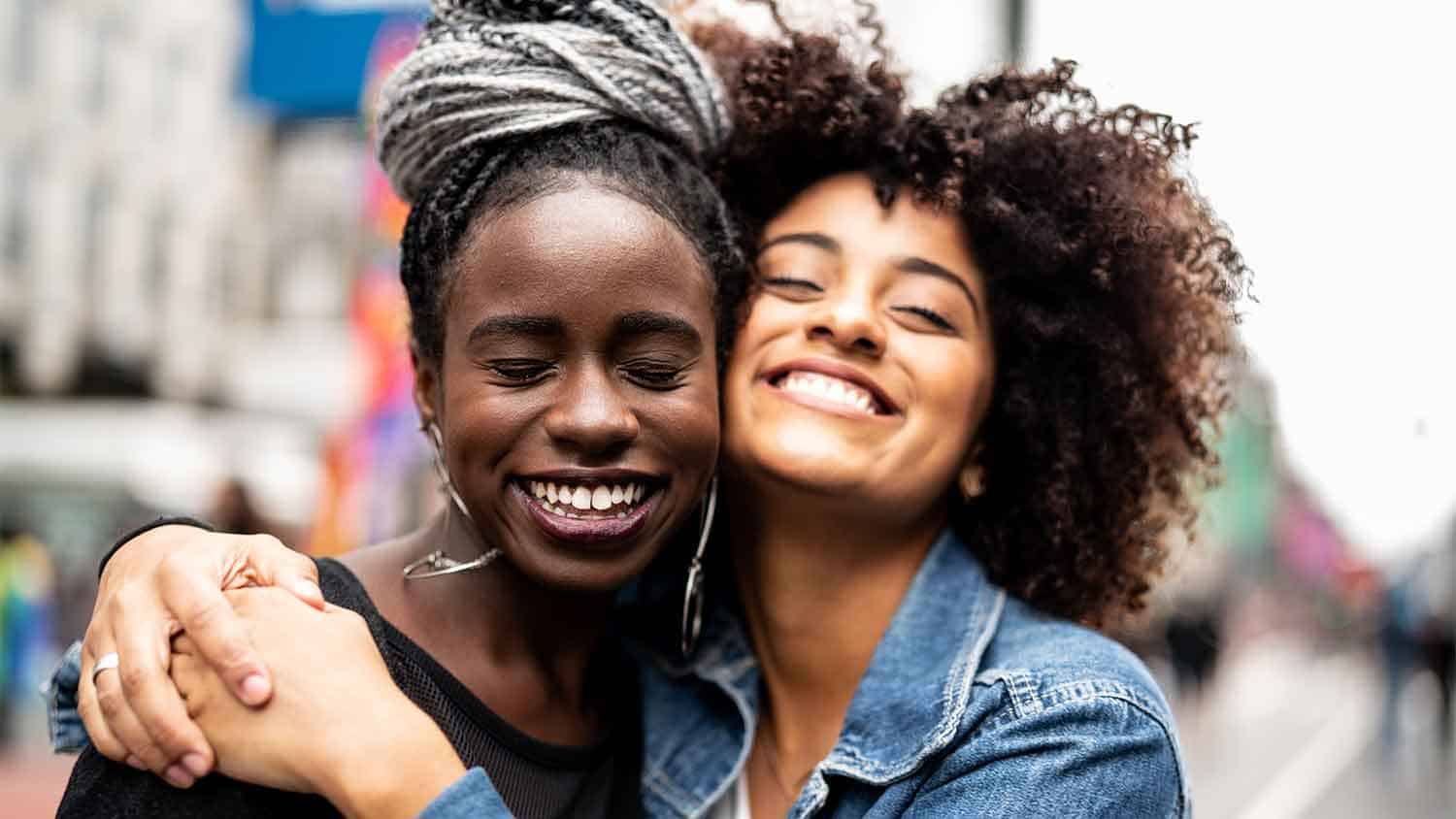 Two women smiling at the camera with eyes closed. They're hugging.