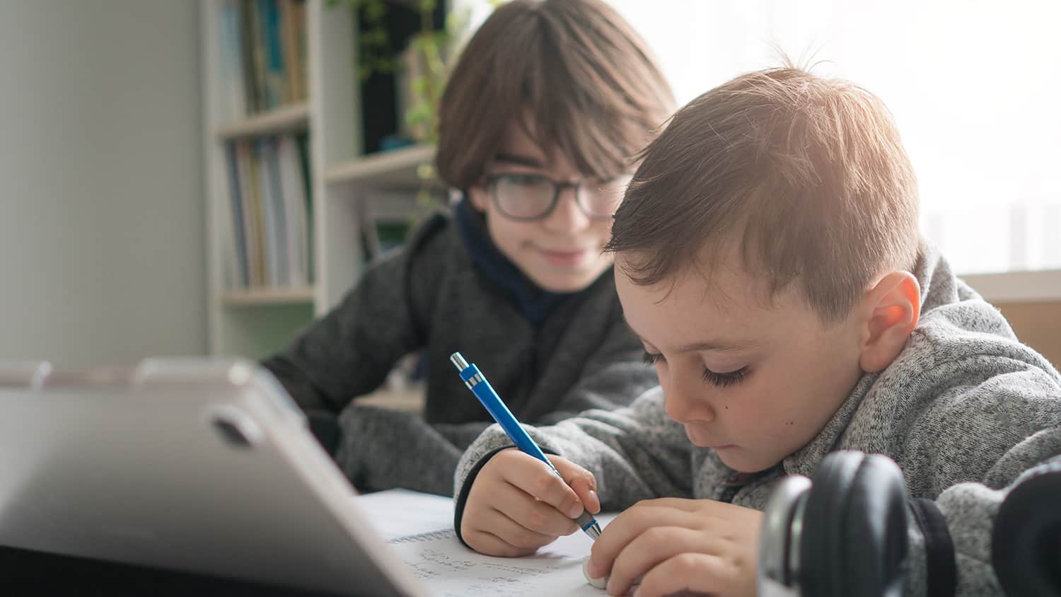 Boy sitting at a desk learning at home using a tablet and notepad, helped by his brother.