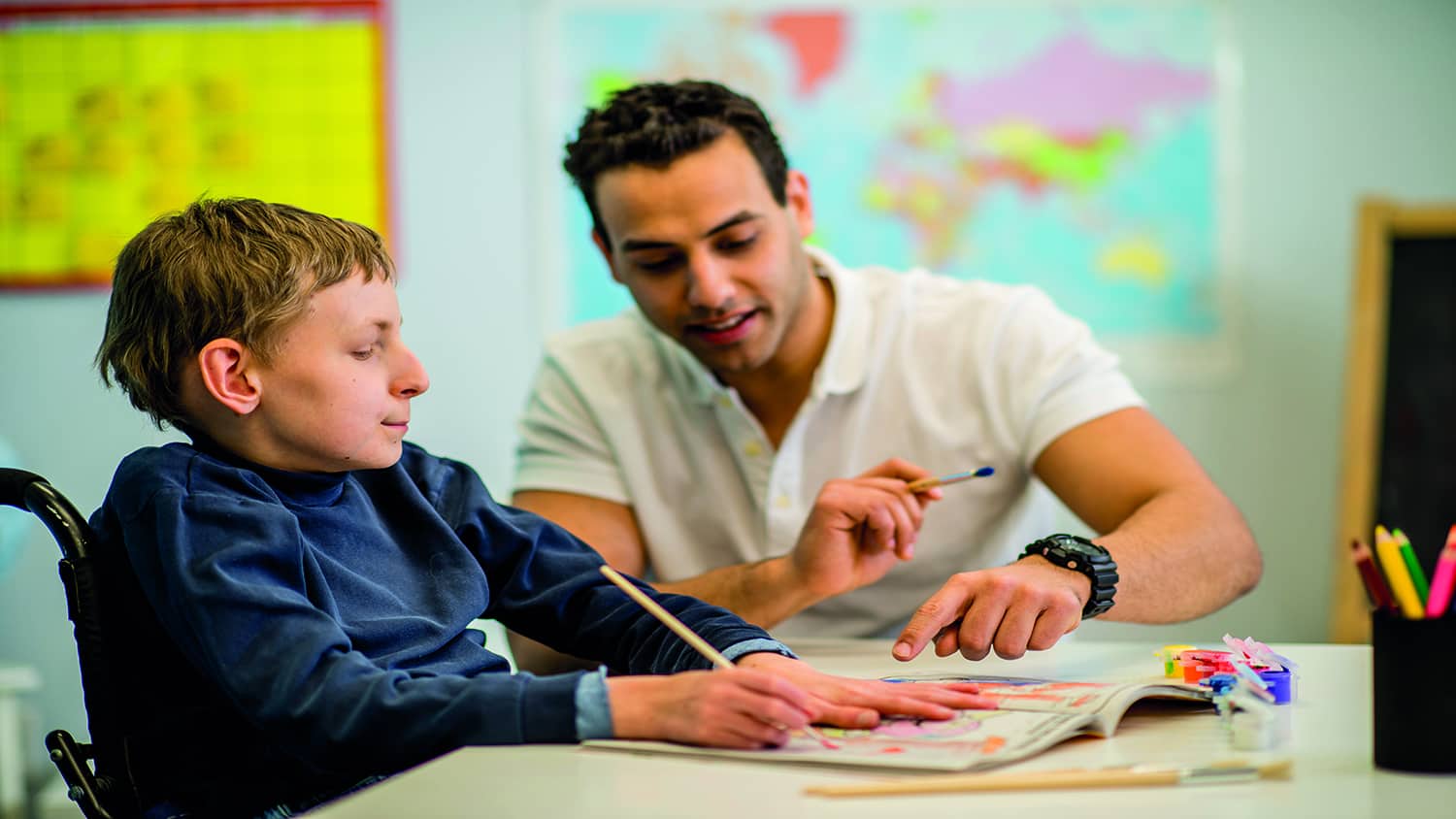 A boy in a wheelchair in a classroom sitting with a teacher