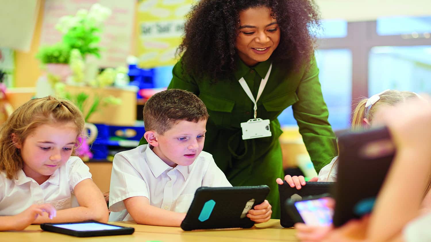 For primary aged schoolchildren sit at their desks looking at digital tablets, with their schoolteacher standing next to them.