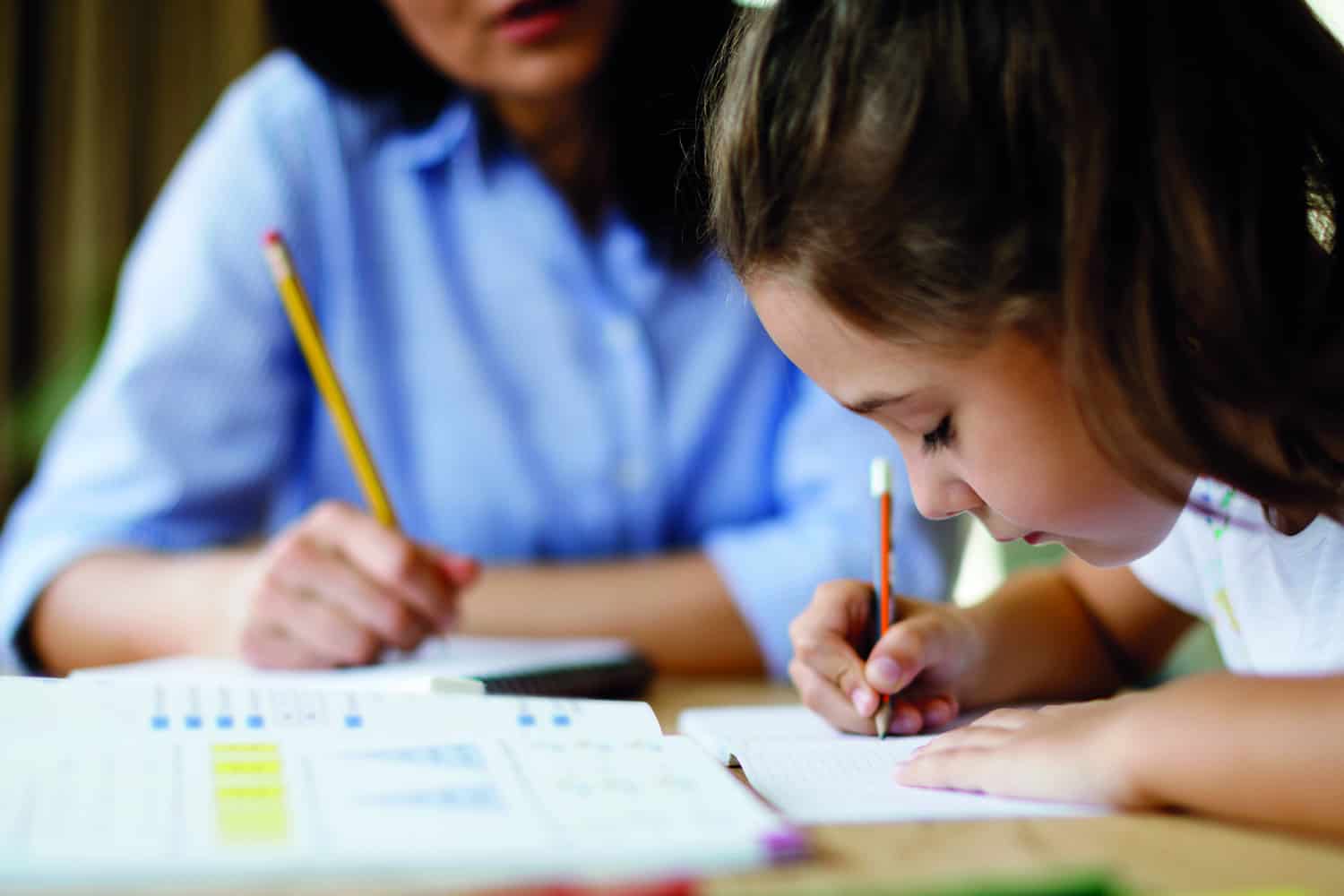 A young girl writing in a book while her teacher looks at her.