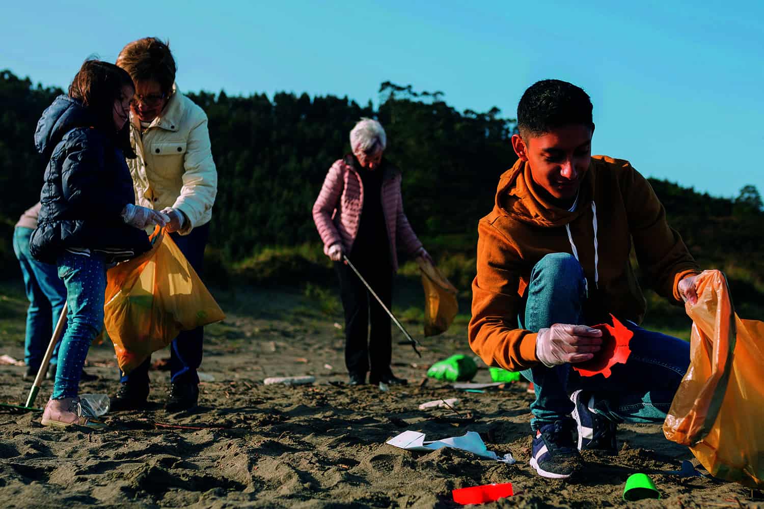 close-up of young person on beach putting litter in a rubbish bag, older woman behind with litter picker and stick. Second pic: woman volunteer handing donations to older woman.