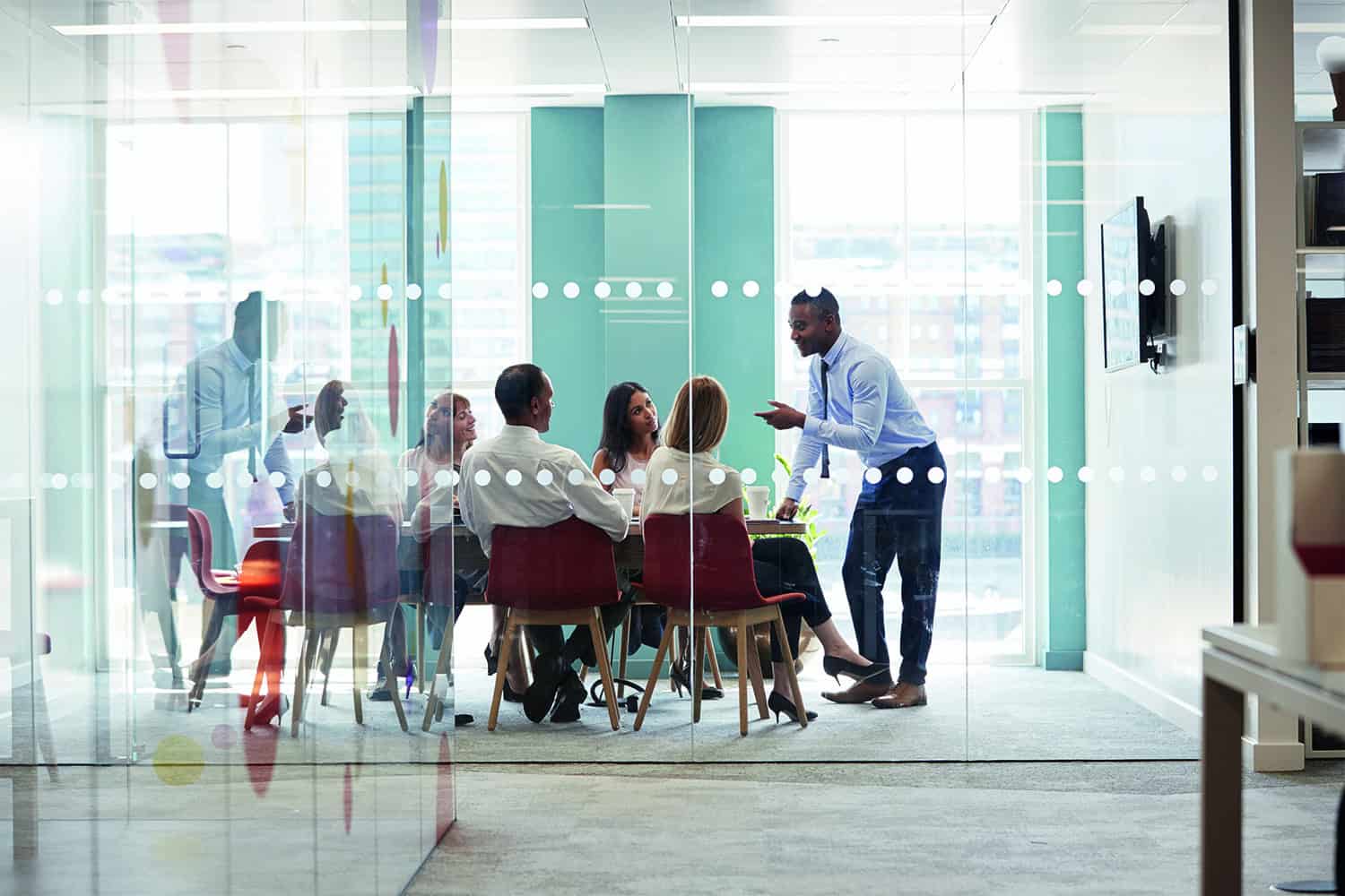 A group of people round a table in an office meeting room with one man standing talking to them.