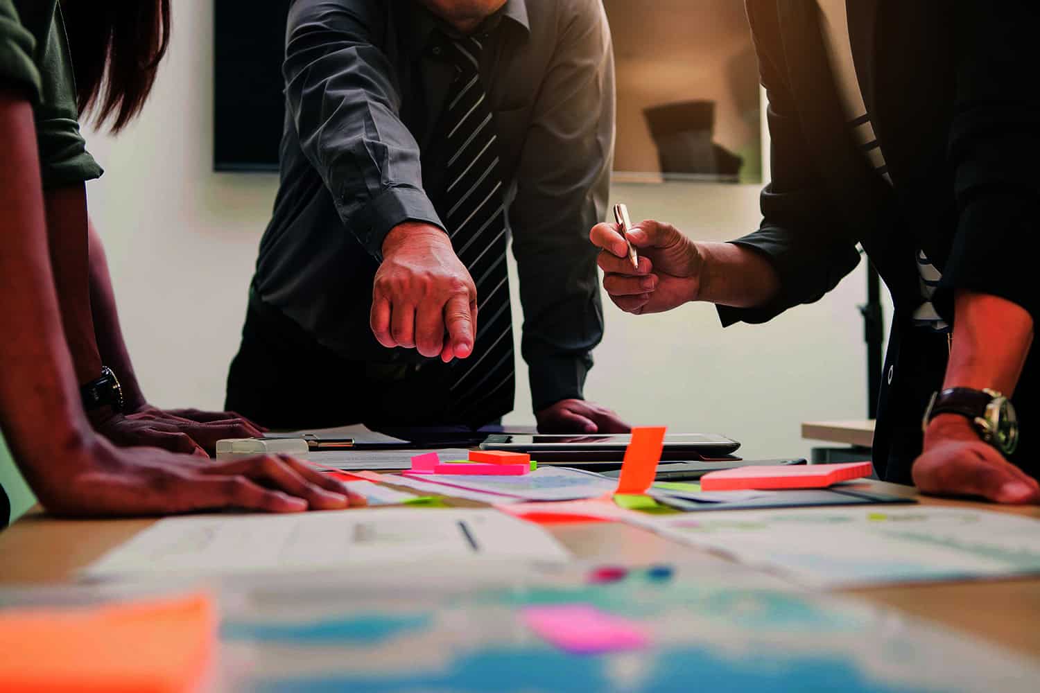 : photo of man in shirt and tie pointing at plans and post-it notes on desk with two others, cropped at chest height.