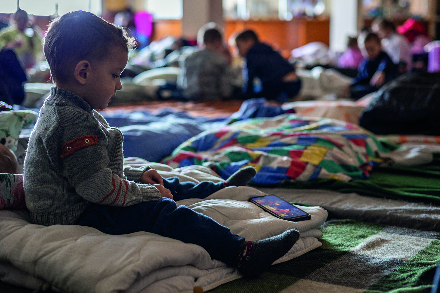 A young boy sitting on his own on a blanket in a large hall looking at a mobile phone