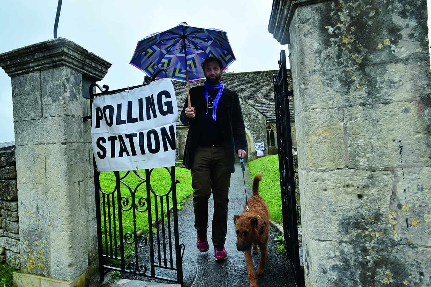 Man with dog and brolly walking out of polling station gate.