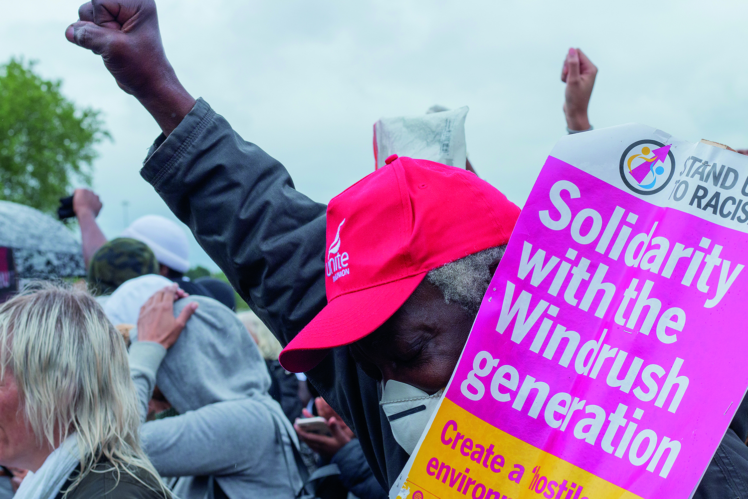 Photo of older black man with fist up carrying 'Solidarity with the Windrush generation' banner in the middle of a crowd of demonstrators.