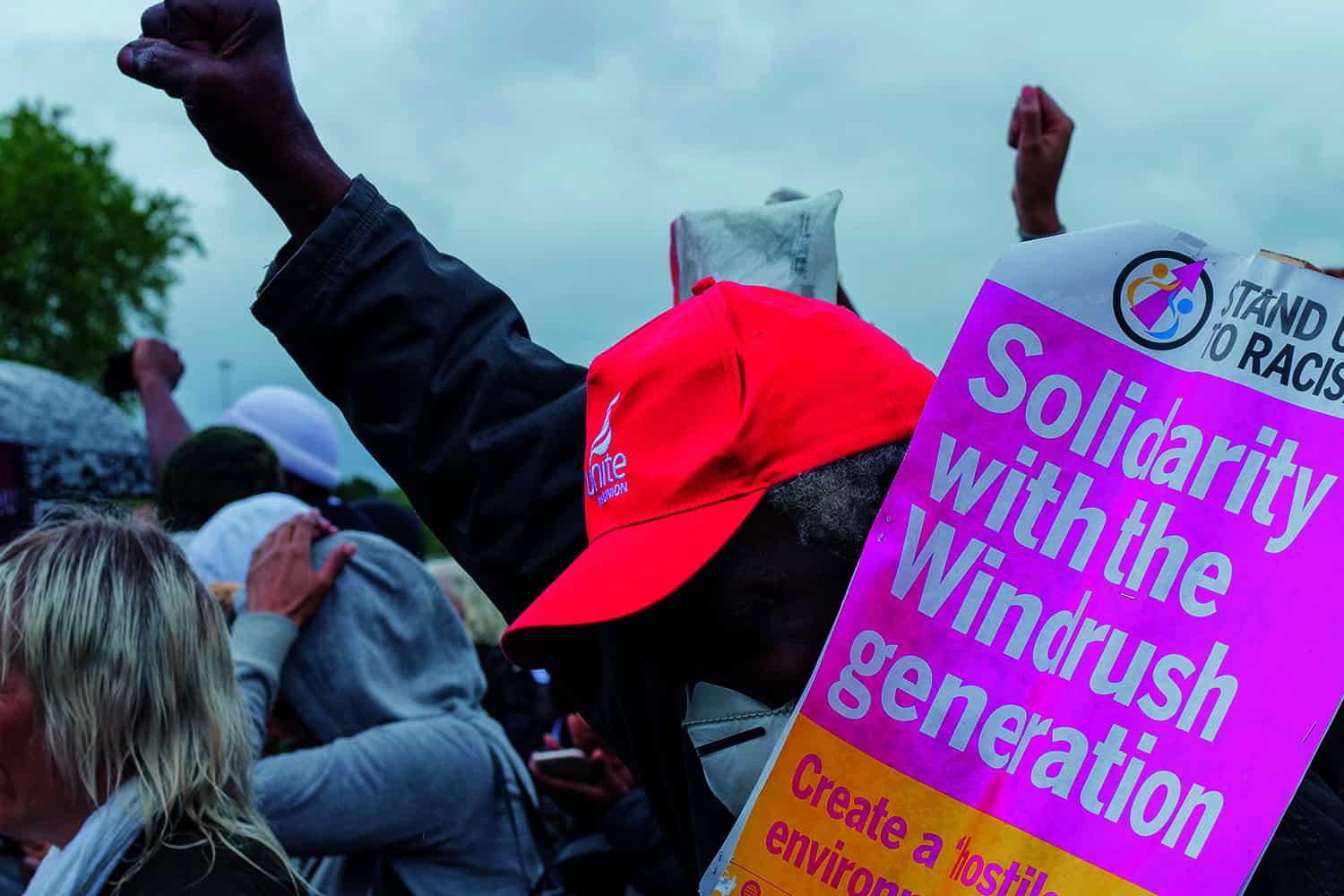 Photo of older black man with fist up carrying 'Solidarity with the Windrush generation' banner in the middle of a crowd of demonstrators.